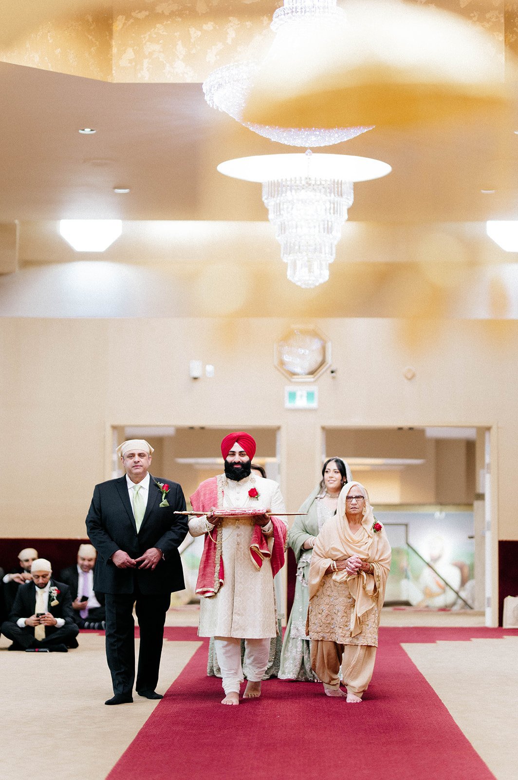 A indian groom in traditional dress is escorted by his parents down the aisle in a gurdwara