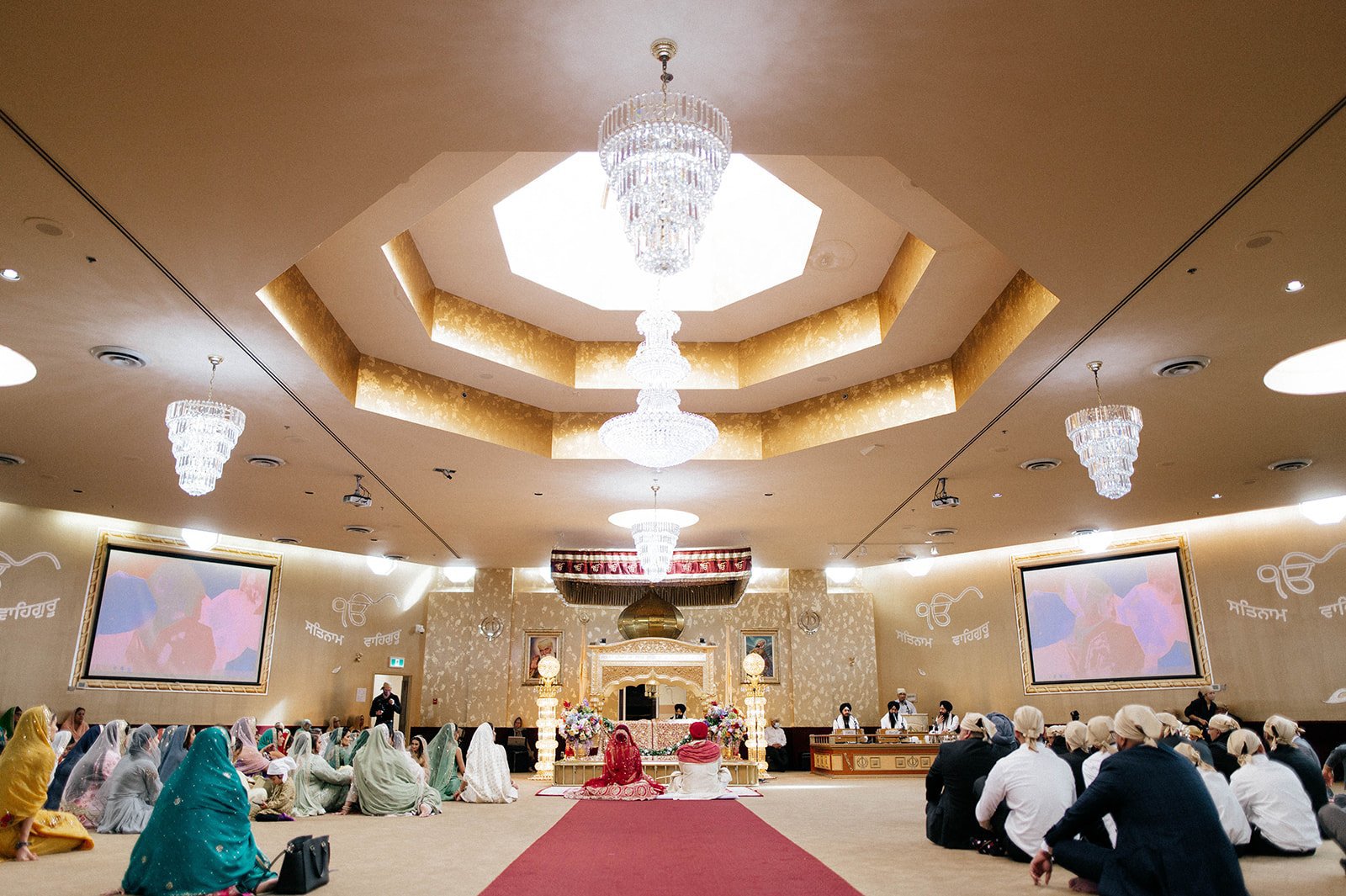 A wide photograph of an indian wedding ceremony in a Gurdwara with a large skylight and chandelier . 