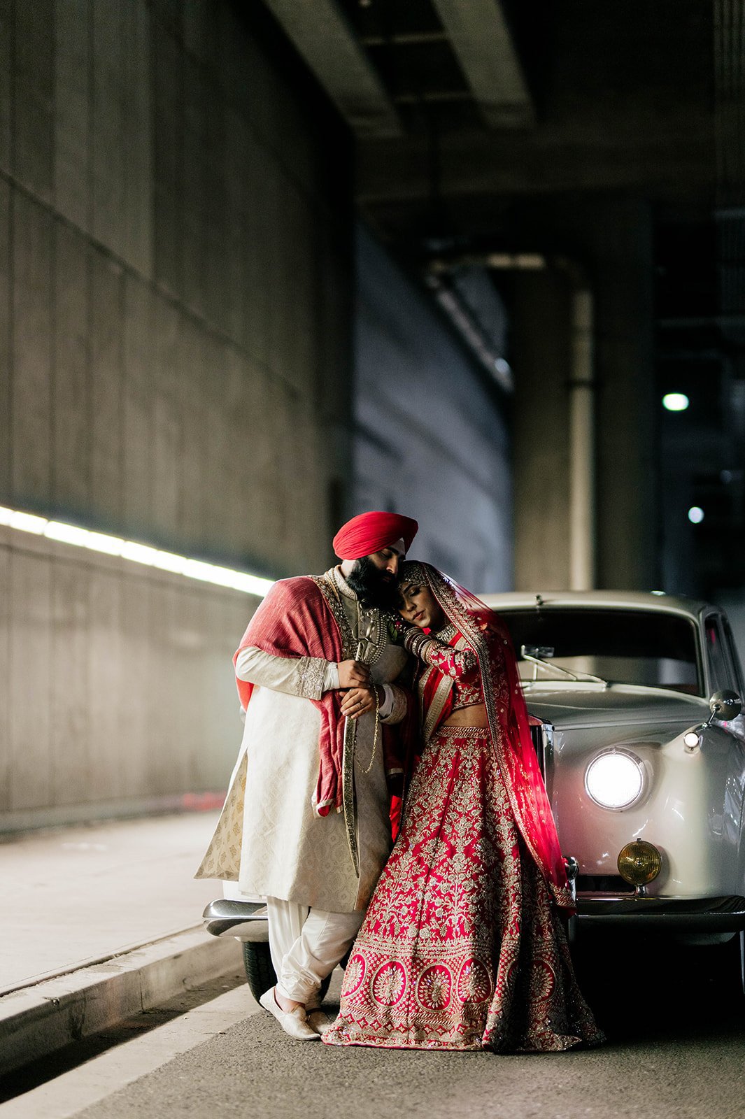 A bride and groom in traditional wedding clothes cuddle on the hood of a vintage car in a surprisingly beautiful tunnel.
