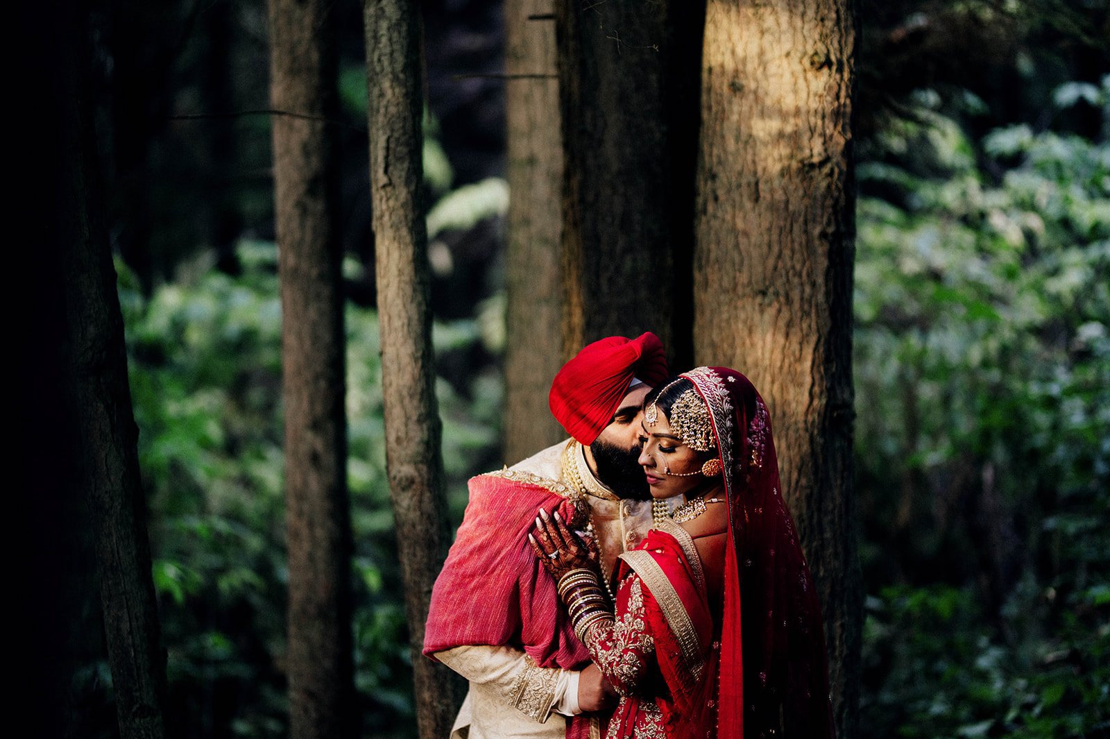 A South asian groom kisses his bride on the cheek in the woods in Stanley Park.