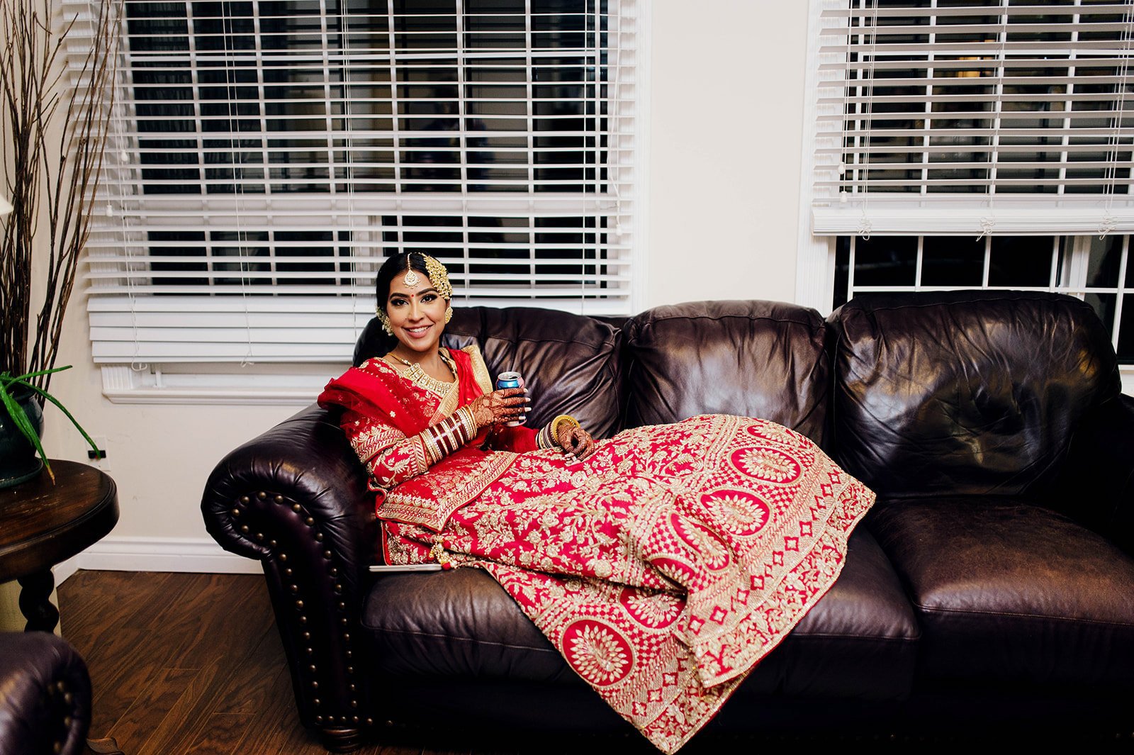 An indian bride relaxes on a brown leather couch as she sips a drink.