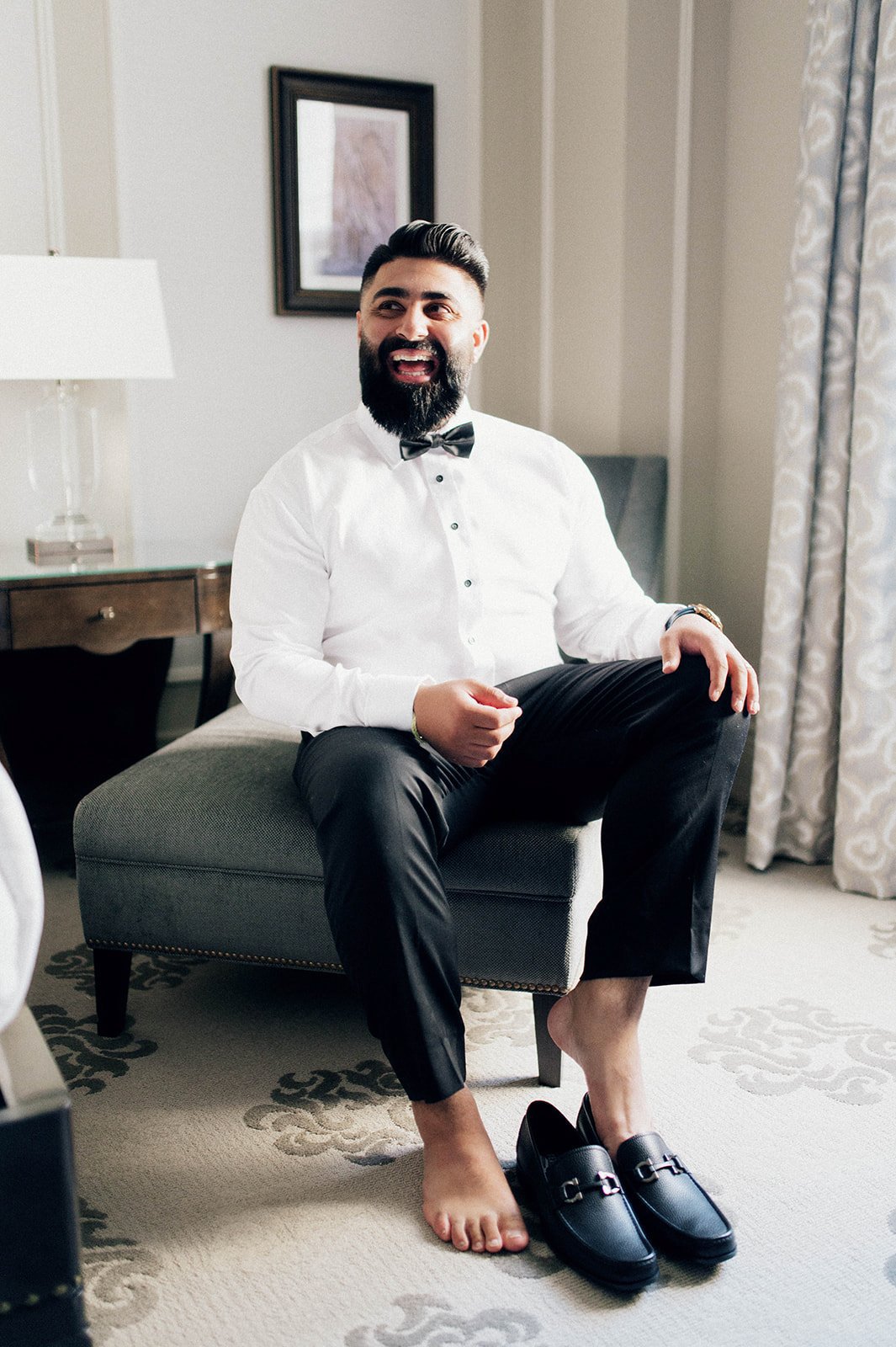 A groom laughs in a hotel room as he puts on his shoes for his reception. 