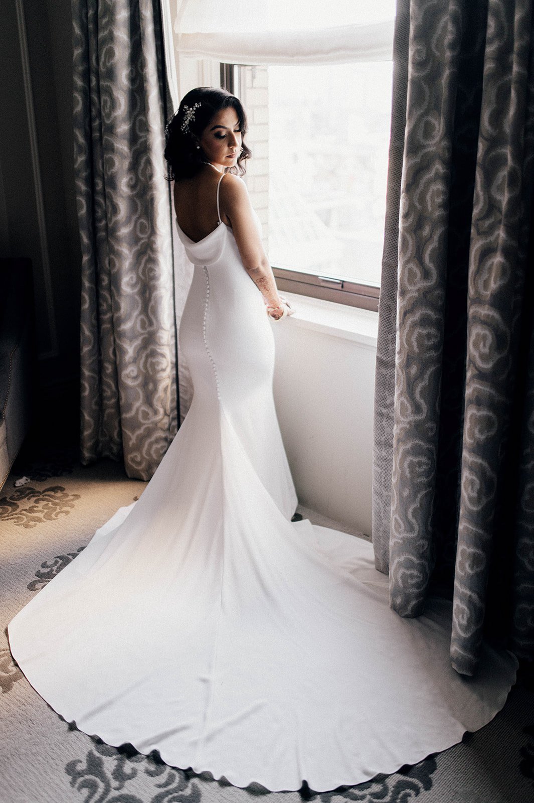 A bride in a long white gown looks over her shoulder as window light streams in.