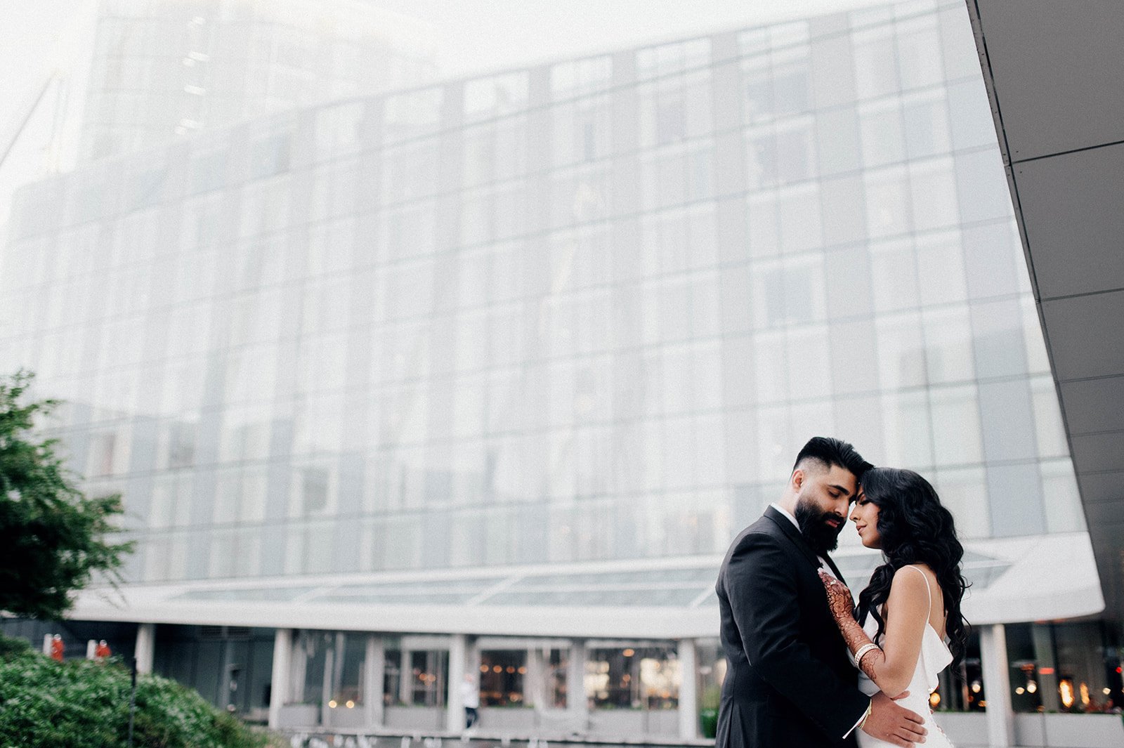 A bride and groom rest their heads together as they are dwarfed by a glass building.