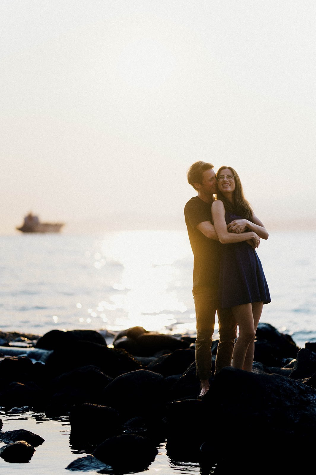 A young couple hug as they stand on the beach during sunset at English Bay, Vancouver, BC. 