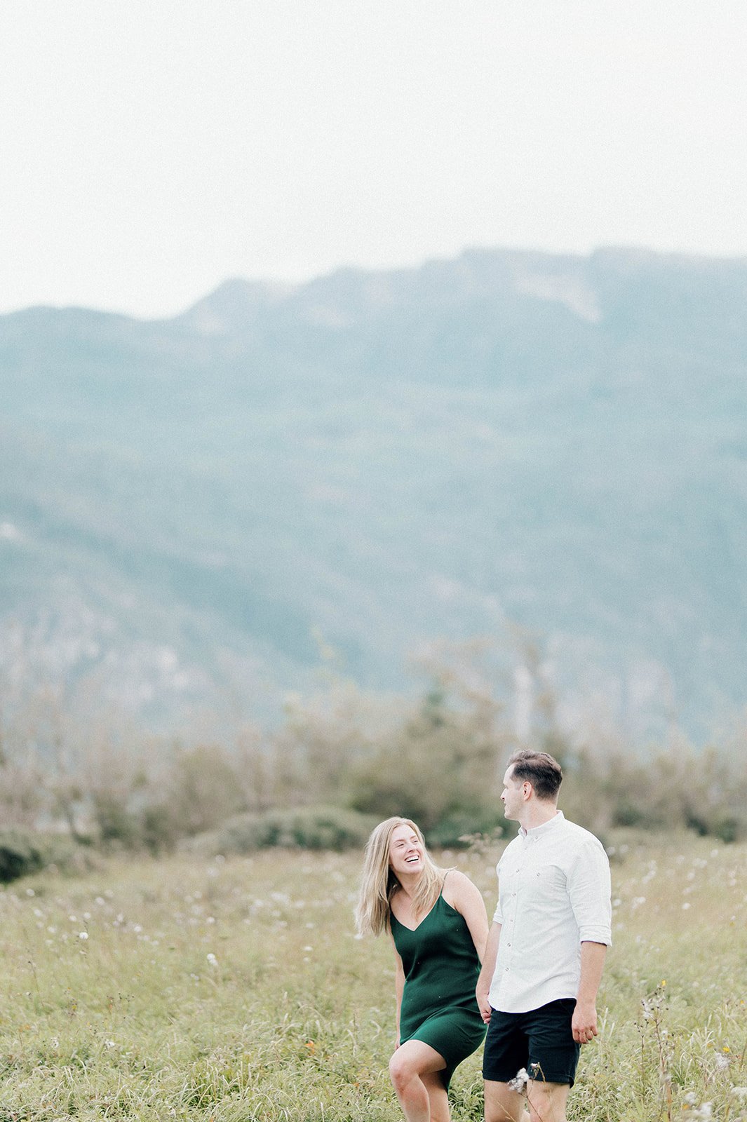 A young and beautiful engaged couple laugh as they stroll through a grassy field in front of a large mountain in Squamish, BC.