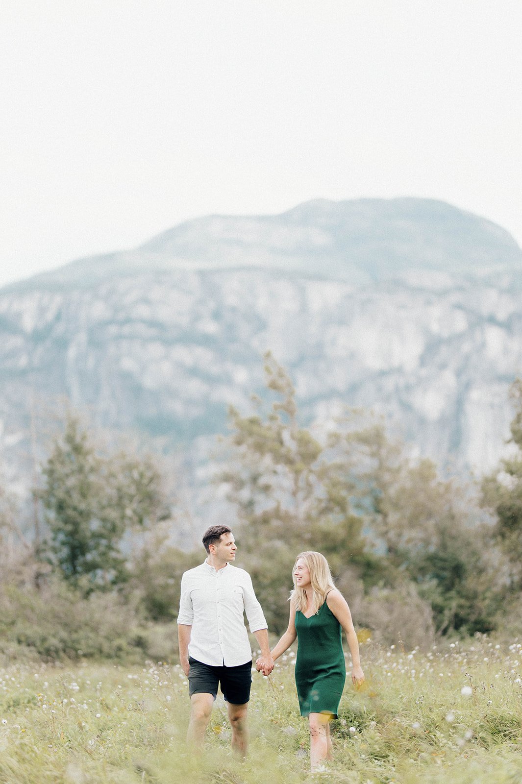 A young and beautiful engaged couple hold hands as they stroll through a grassy field in front of a large mountain in Squamish, BC.