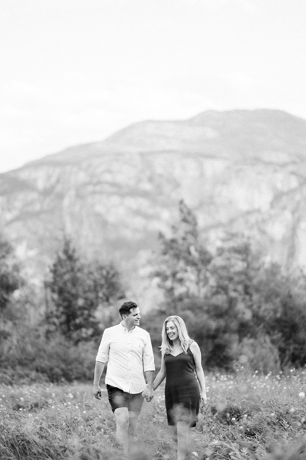 A young and beautiful engaged couple strolls through a grassy field in front of a large mountain in Squamish, BC.