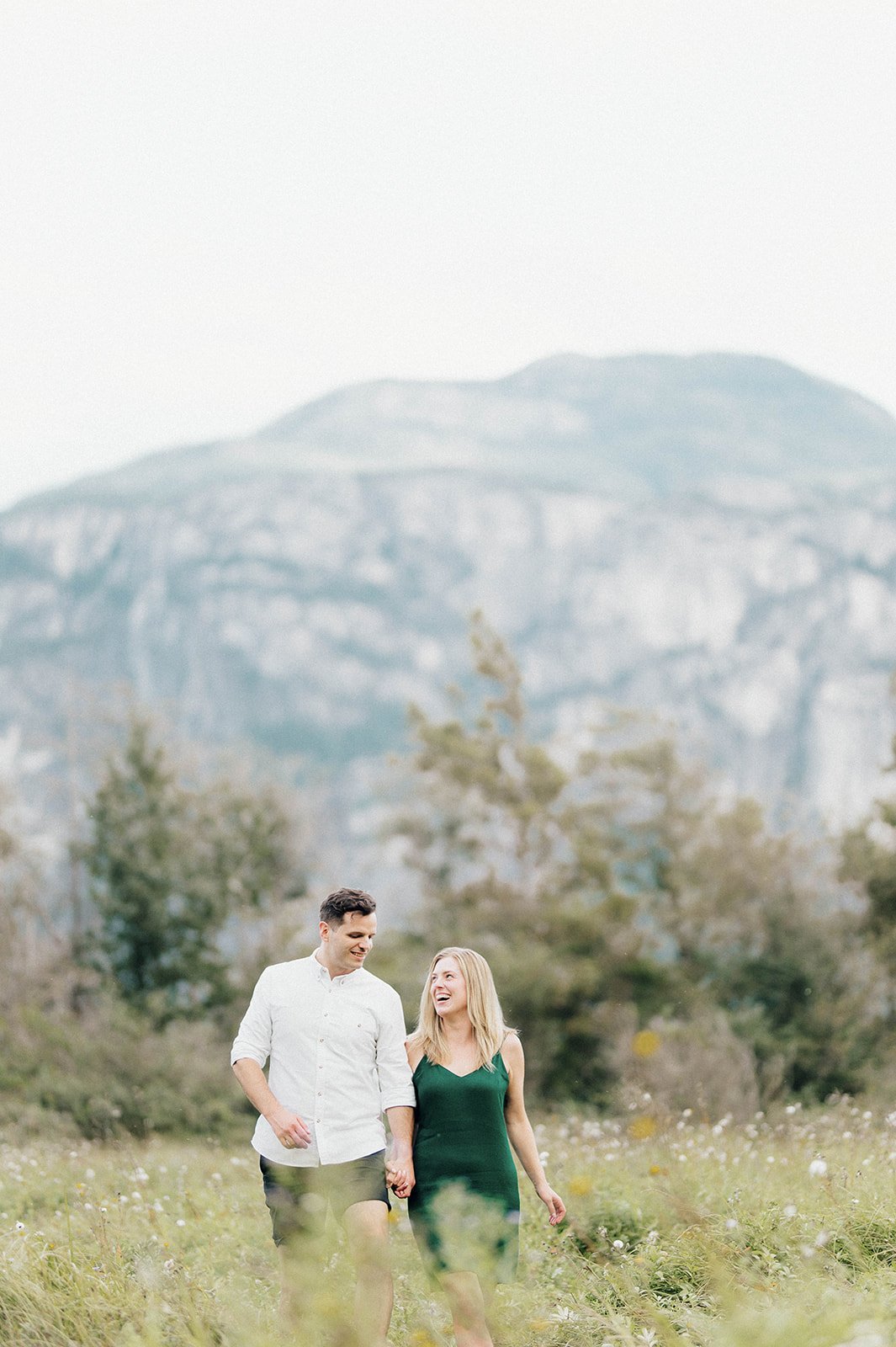 A young and beautiful engaged couple strolls through a grassy field in front of a large mountain in Squamish, BC.