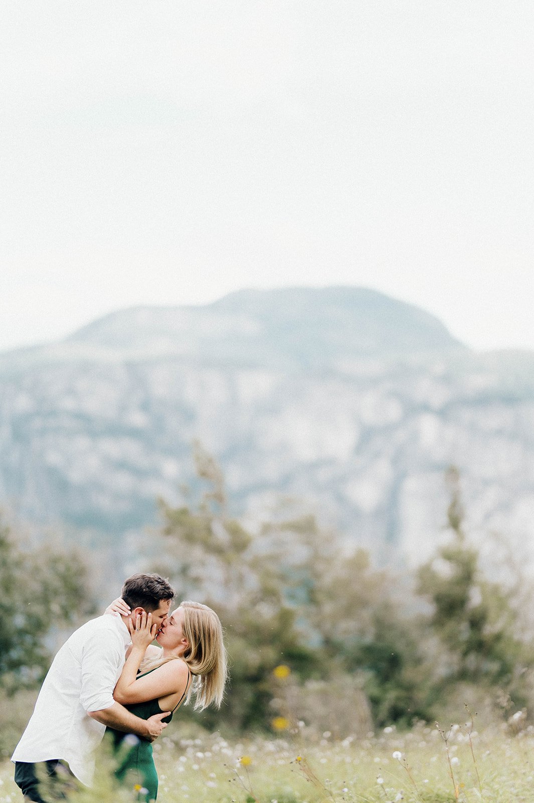 A young and beautiful engaged couple passionately kiss in a grassy field in front of a large mountain in Squamish, BC.