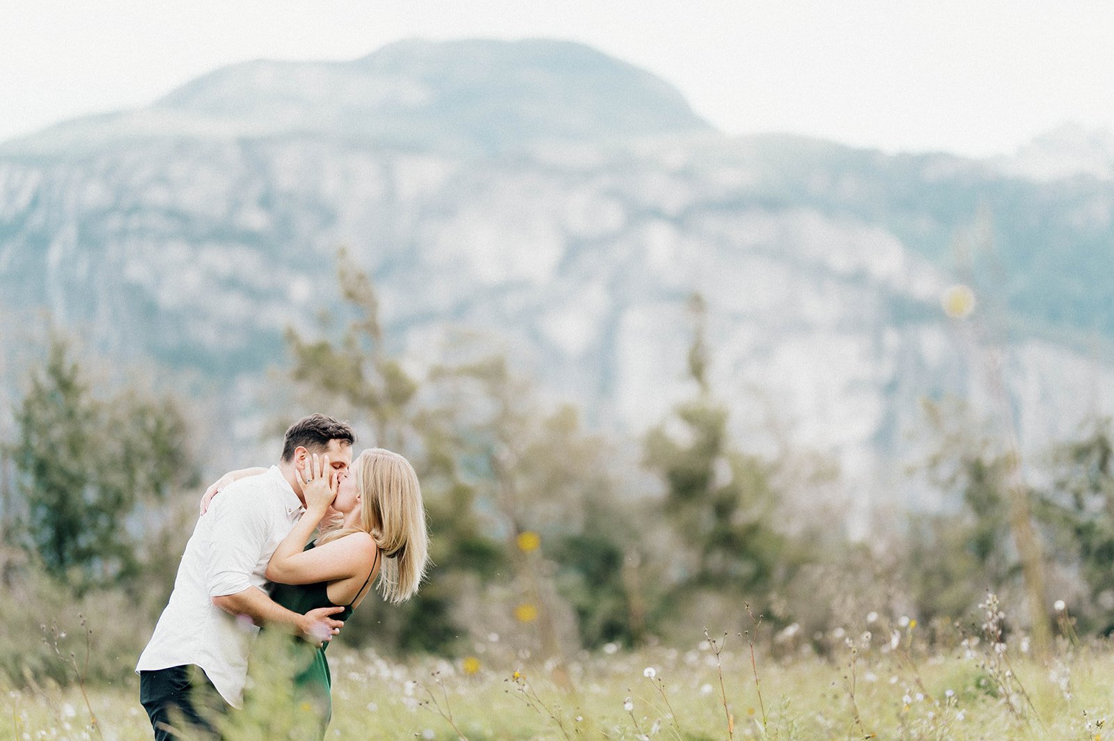 A young and beautiful engaged couple passionately kiss in a grassy field in front of a large mountain in Squamish, BC.