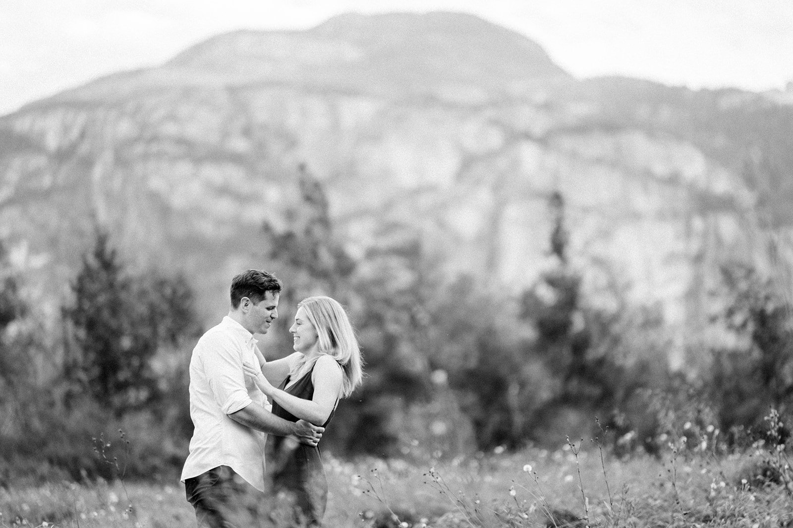 A young and beautiful engaged couple canoodle in a grassy field in front of a large mountain in Squamish, BC.