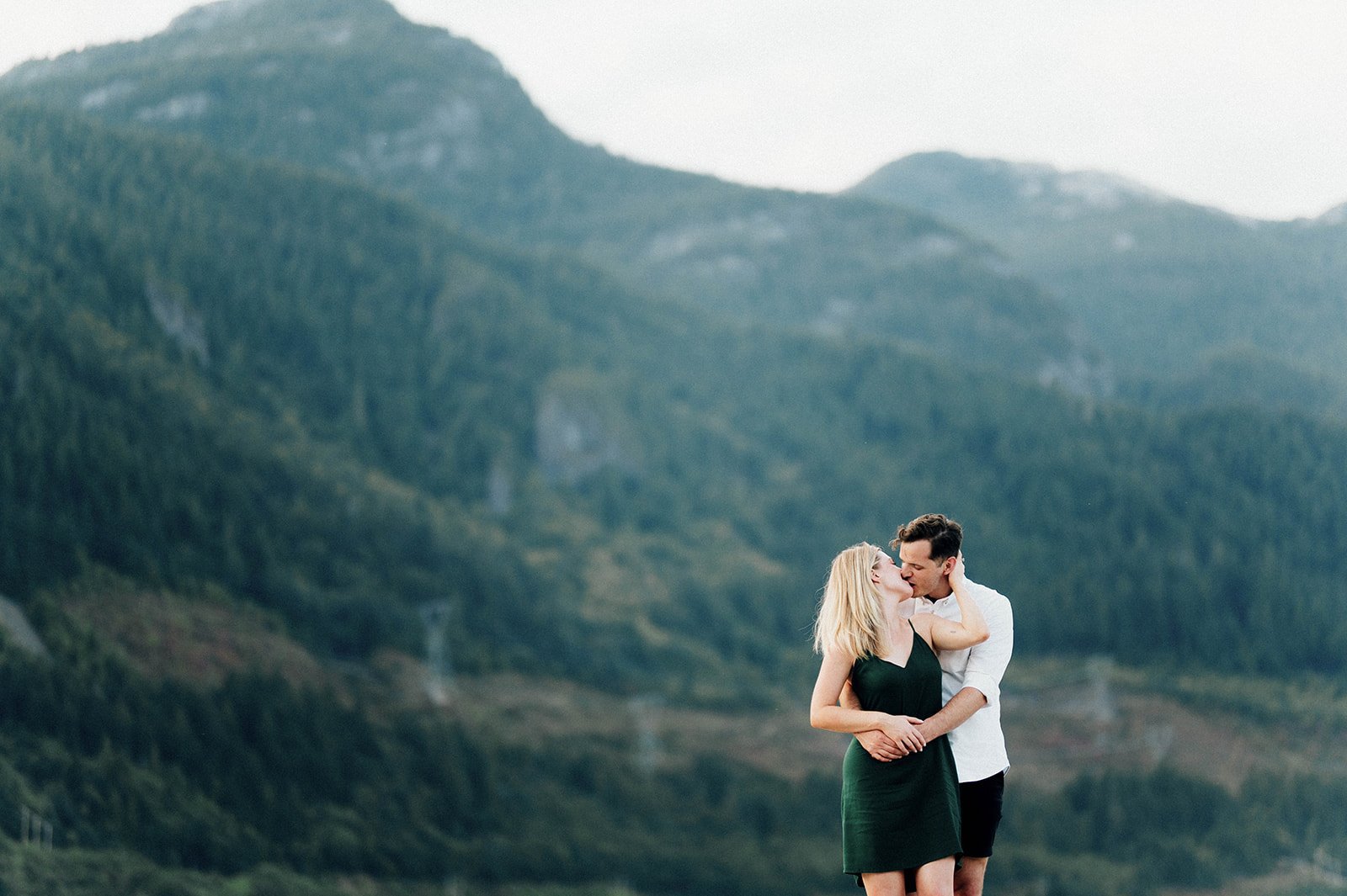 A man lovingly embraces his fiancee from behind as she kisses him in front of and emerald green mountain.