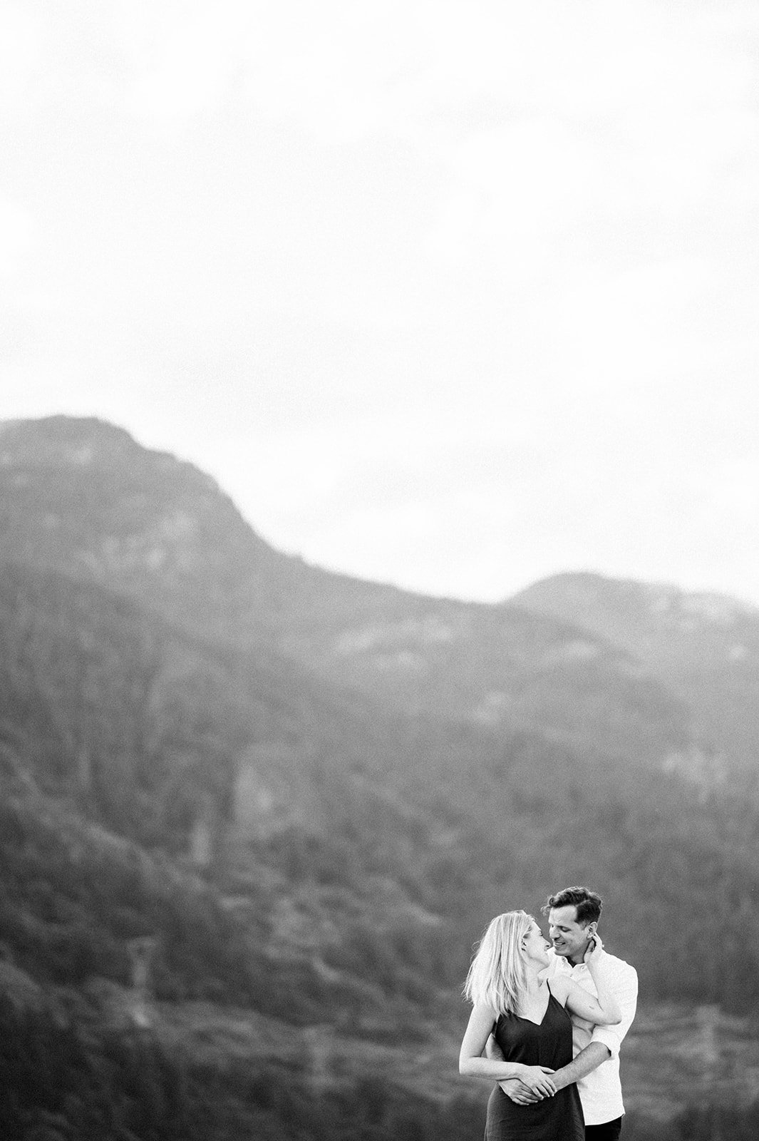 A man lovingly embraces his fiancee from behind as she stroked his hair in front of and emerald green mountain.