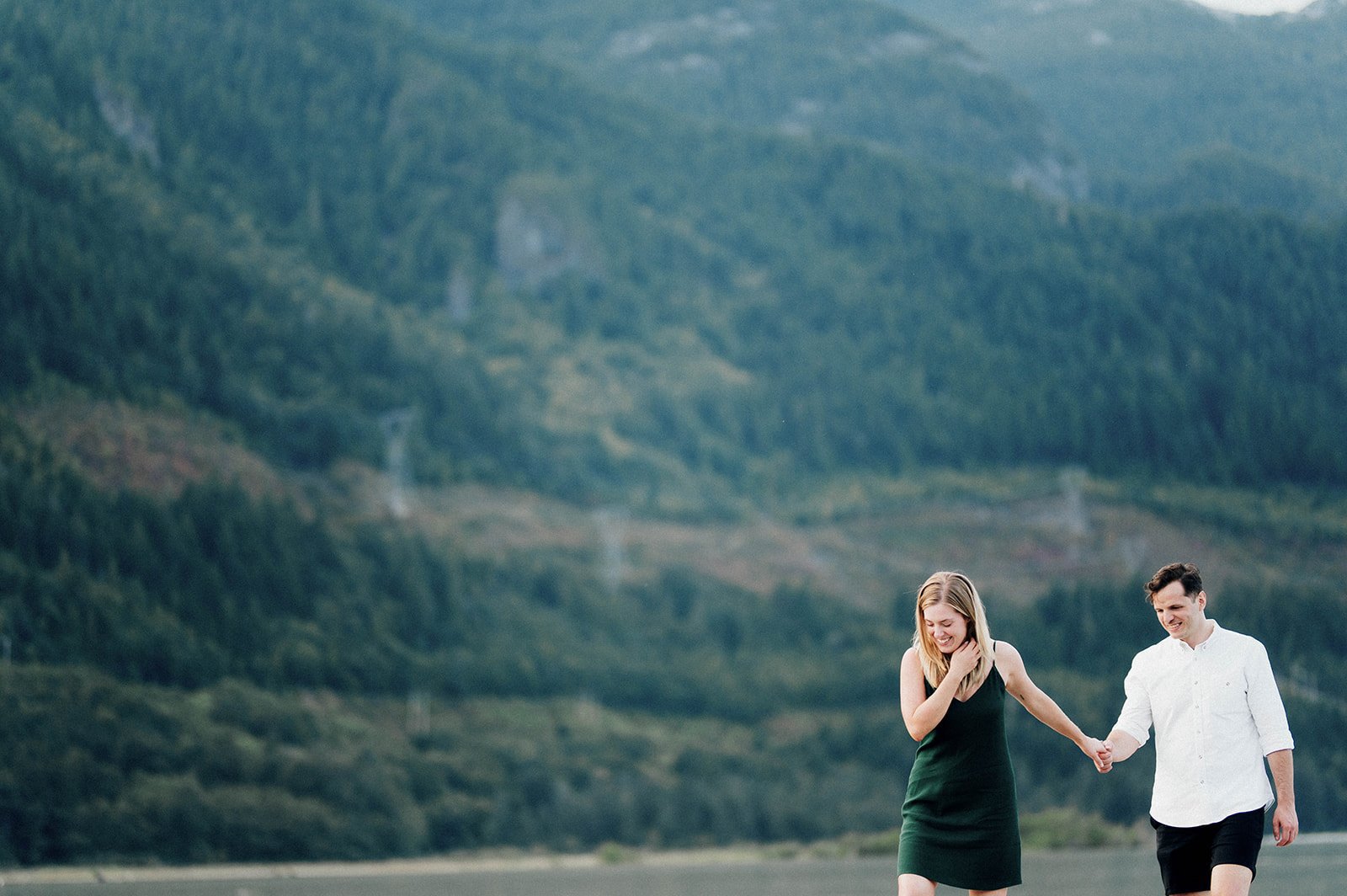 A blonde woman leads a man as they walk through a field in front of the mountains of Squamish, BC.