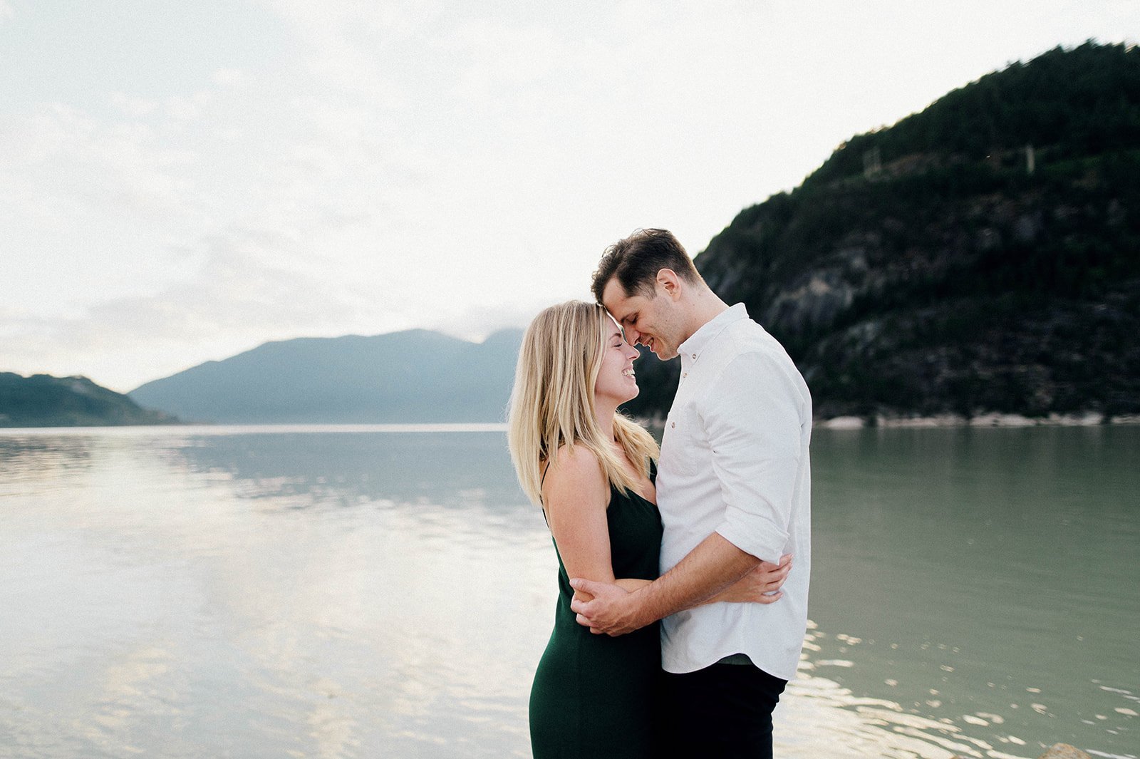 A man and woman embrace in front of a scenic lake surrounded by mountains in Squamish, BC    