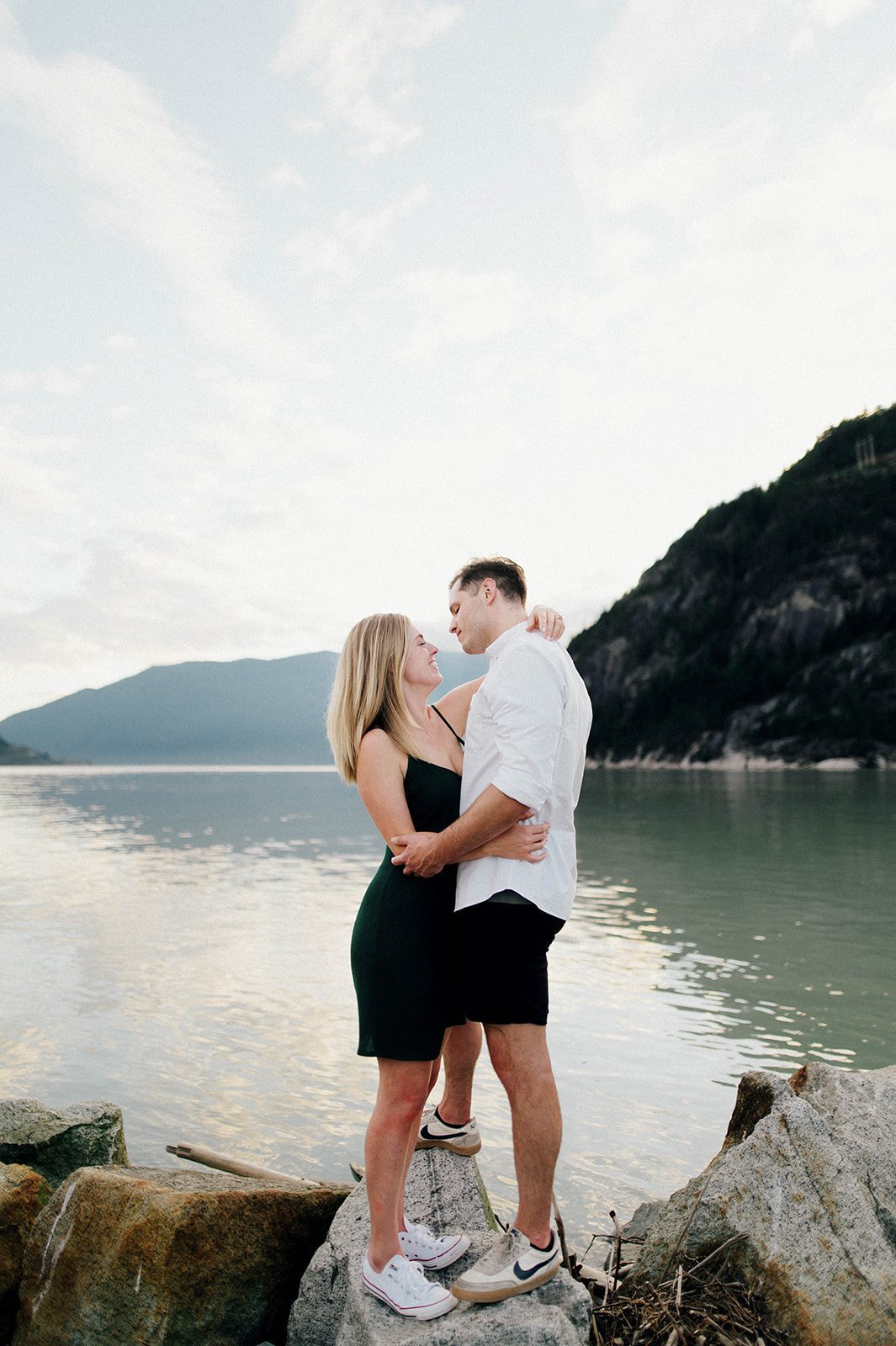 A man and woman embrace in front of a scenic lake as they stand on rocks,surrounded by mountains in Squamish, BC   