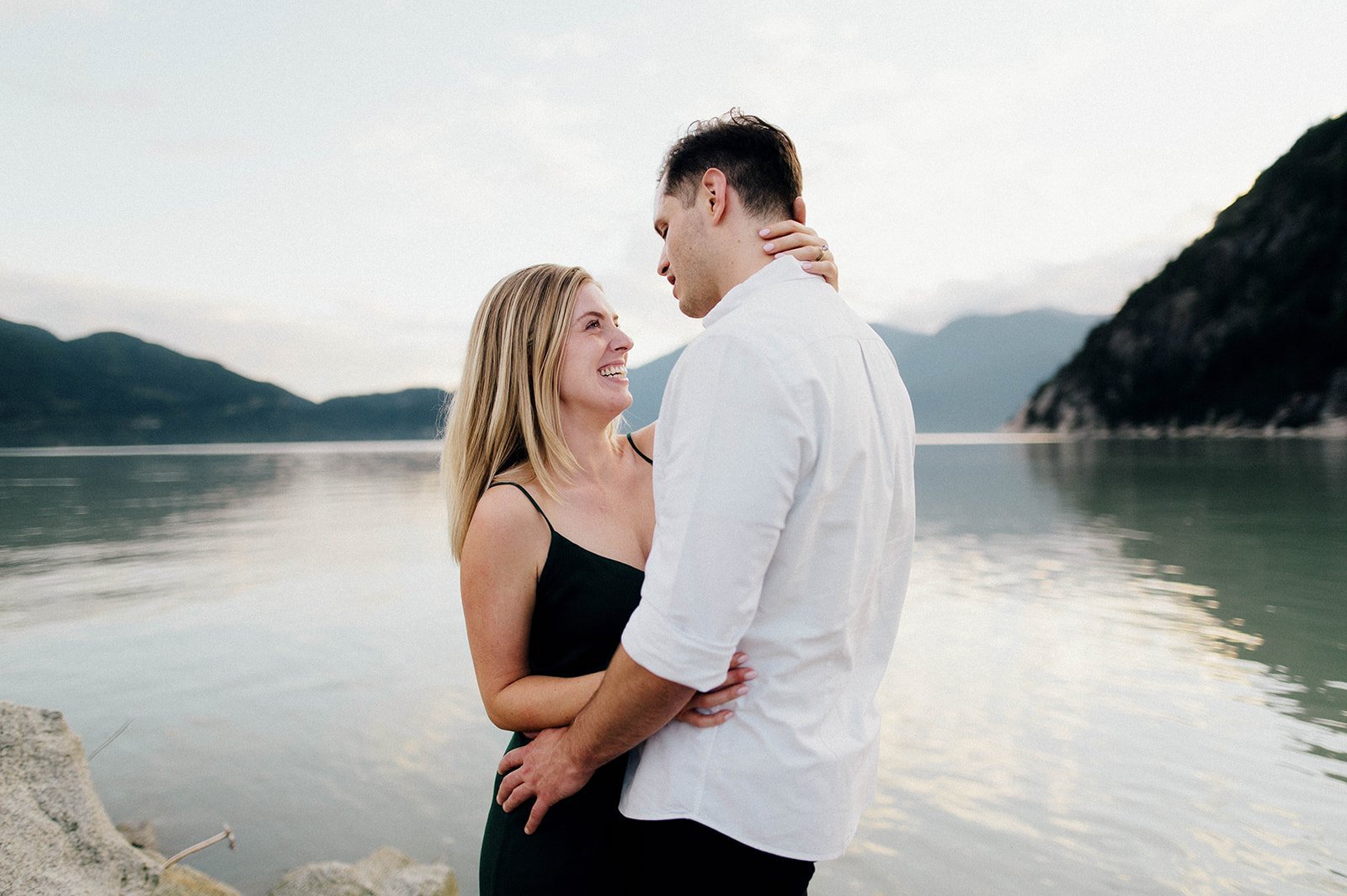 A man and woman embrace and smile at each other in front of a scenic lake surrounded by mountains in Squamish, BC   