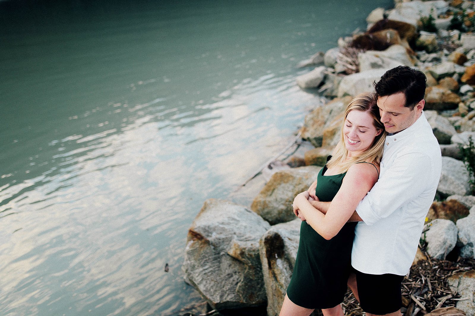 A man and woman in an green dress embrace in front of a scenic lake surrounded by mountains in Squamish, BC   