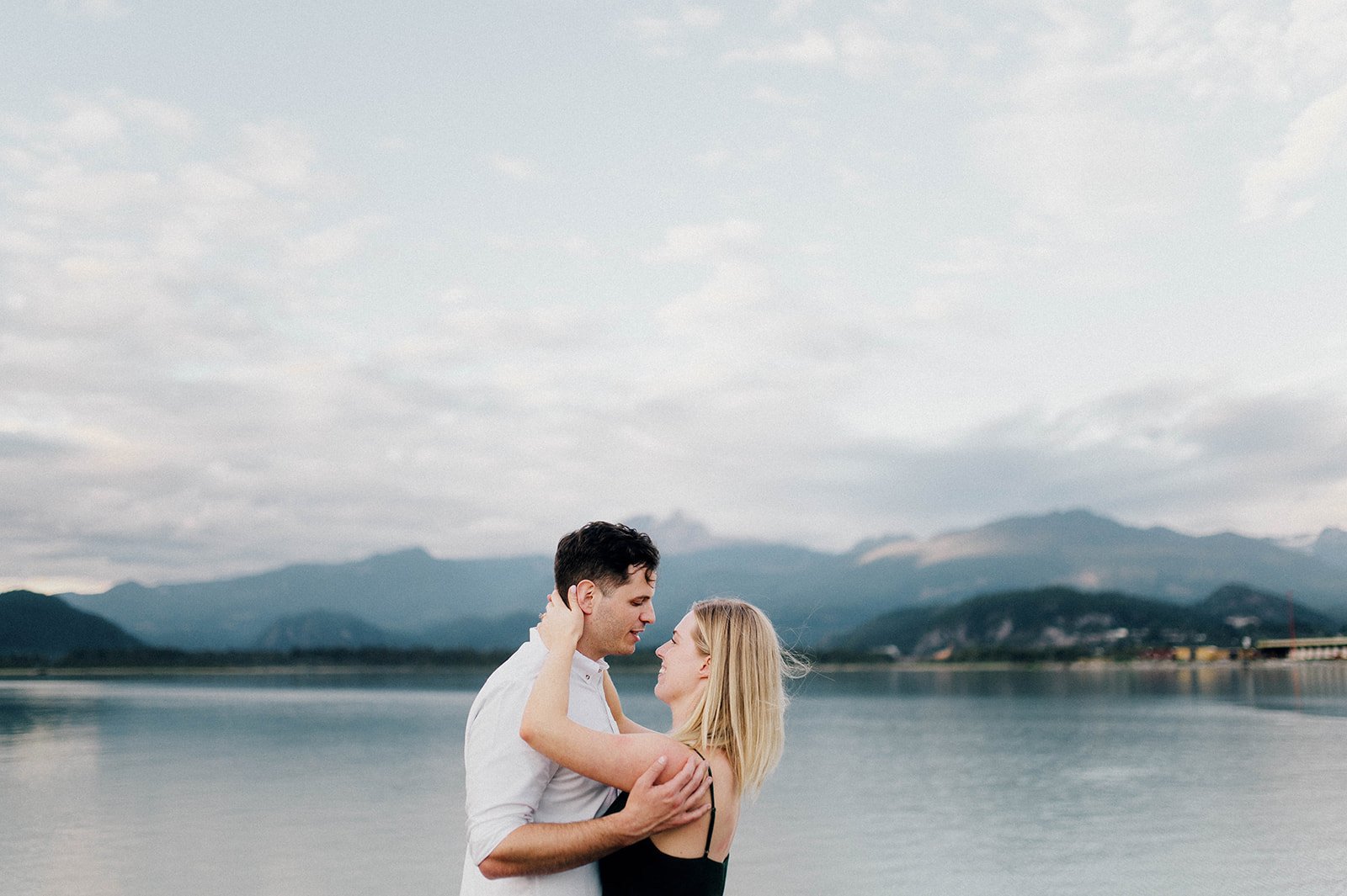 A man and woman embrace in front of a scenic lake surrounded by mountains in Squamish, BC   