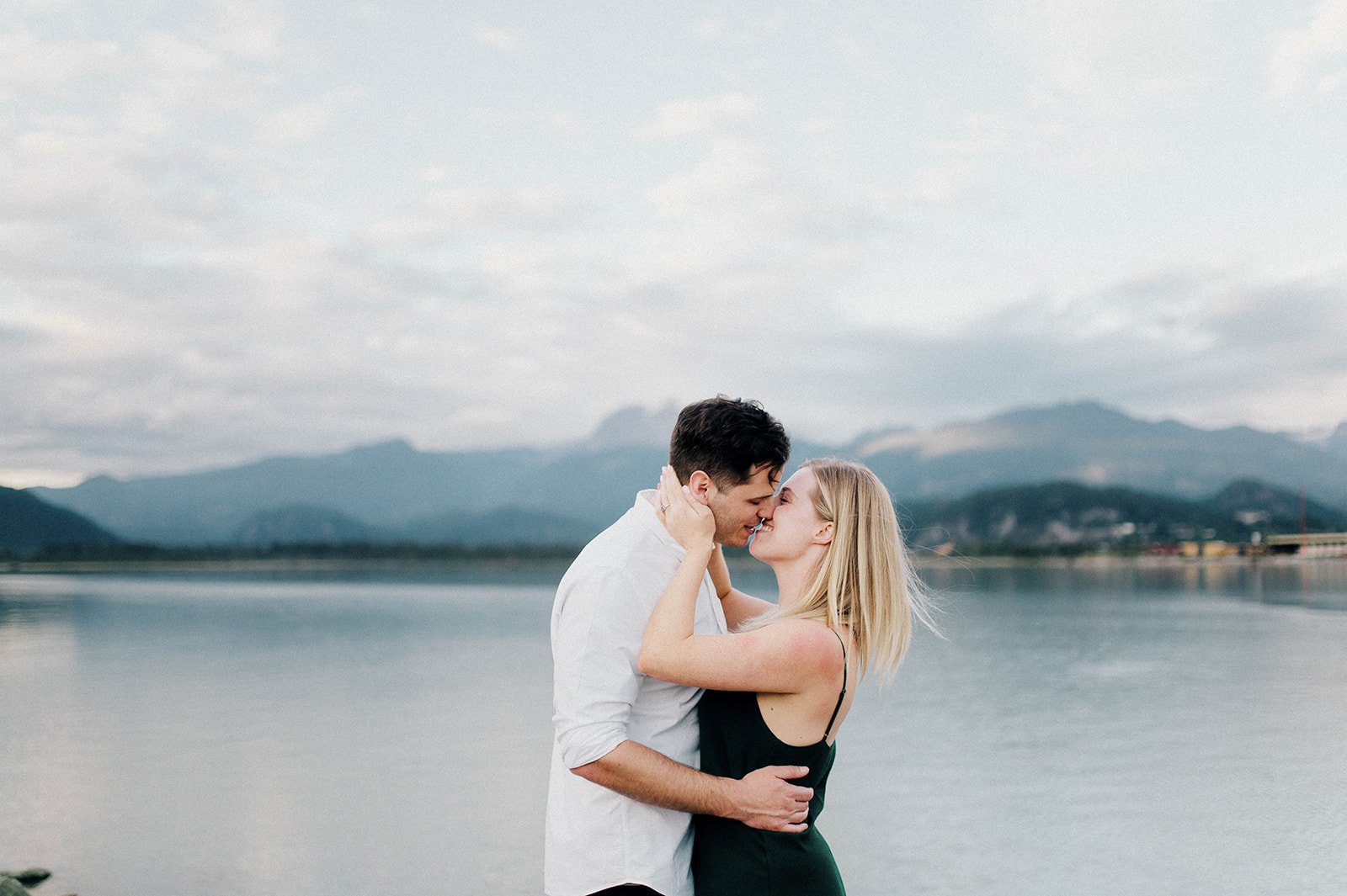 A man and woman share a kiss in front of a scenic lake surrounded by mountains in Squamish, BC   