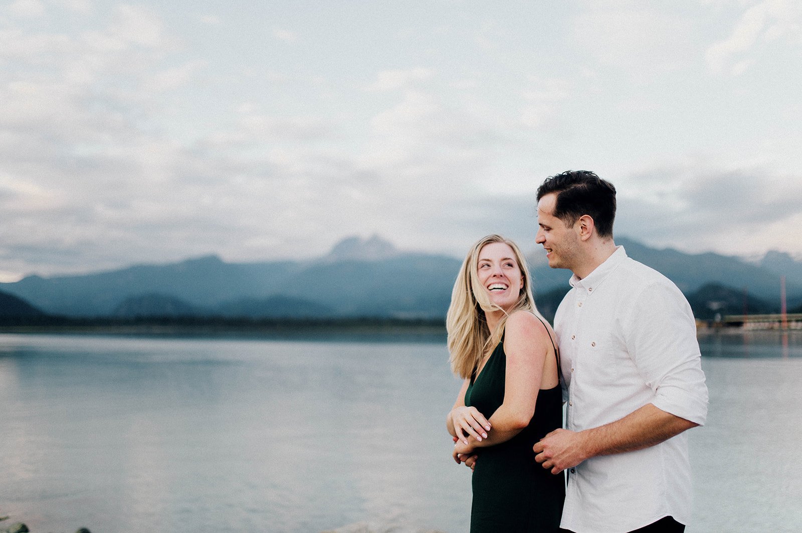 A man and woman laugh as they cuddle in front of a scenic lake surrounded by mountains in Squamish, BC   