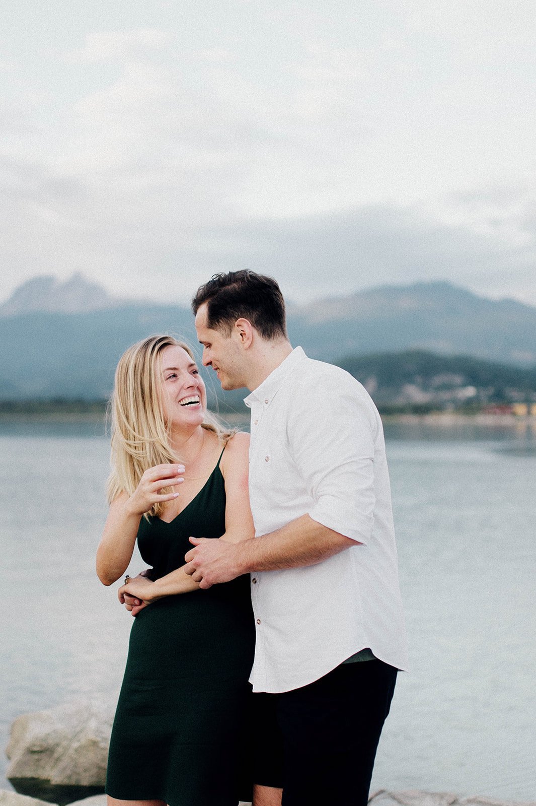 A man and woman chuckle as they hug in front of a scenic lake surrounded by mountains in Squamish, BC   