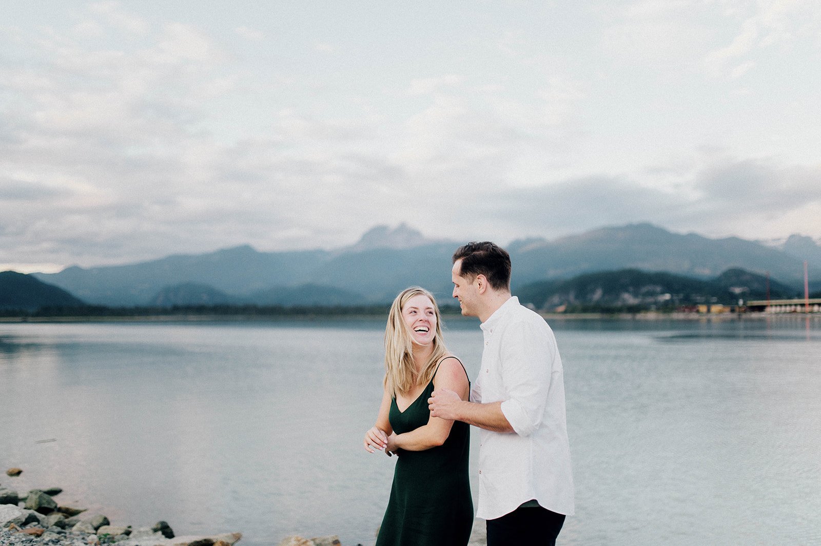 A man and woman embrace in front of a scenic lake surrounded by mountains in Squamish, BC   