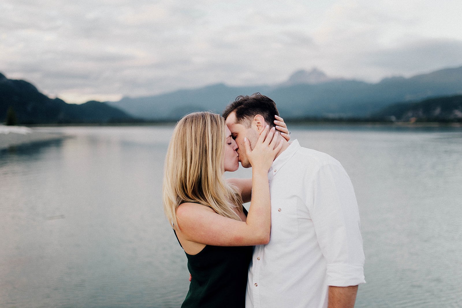 A man and woman kiss in front of a scenic lake surrounded by mountains in Squamish, BC   
