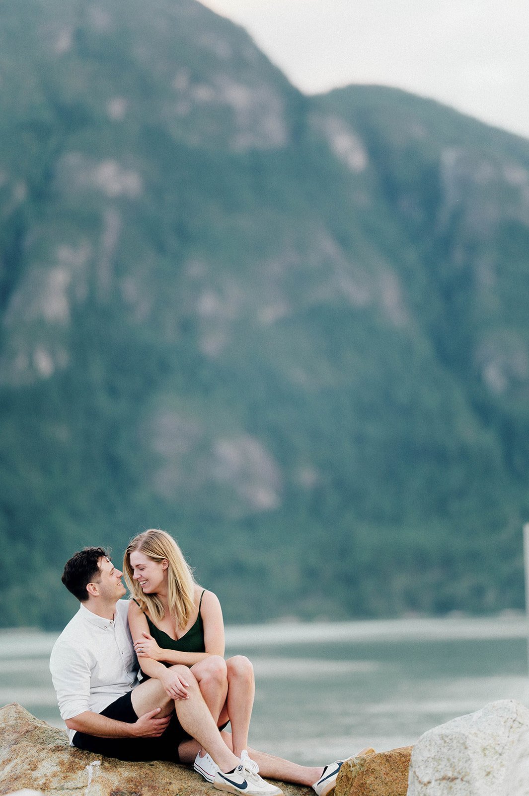 A blonde woman in a green dress sits on a man's lap in front of a beautiful green lake and mountain in Squamish, BC.