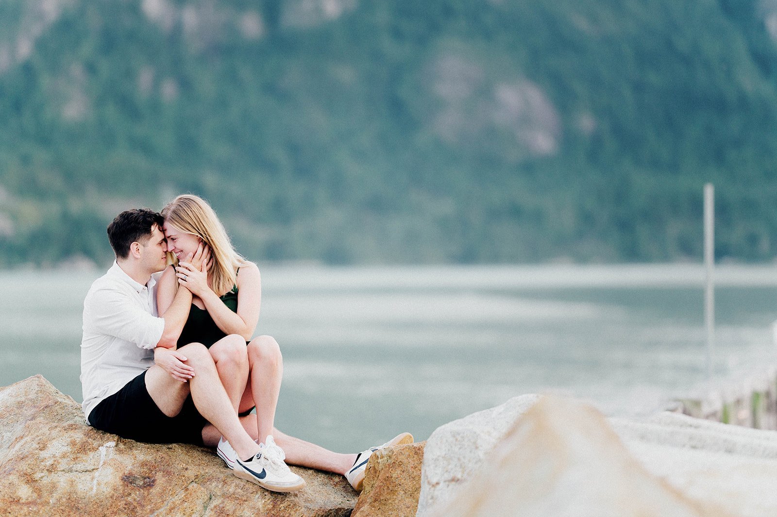A blonde woman in a green dress sits on a man's lap in front of a beautiful green lake and mountain in Squamish, BC.