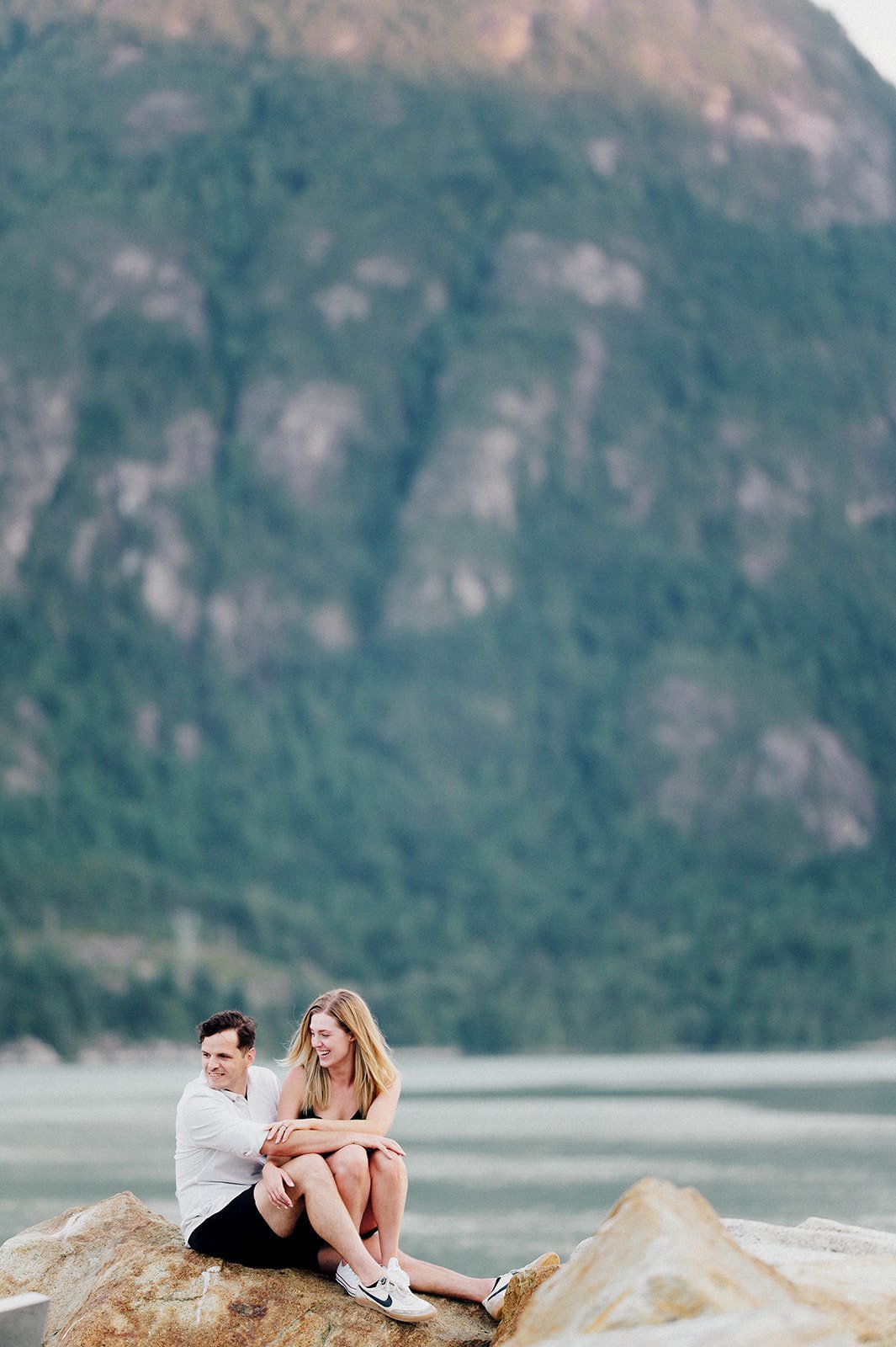 A blonde woman in a green dress sits on a man's lap and laughs in front of a beautiful green lake and mountain in Squamish, BC.