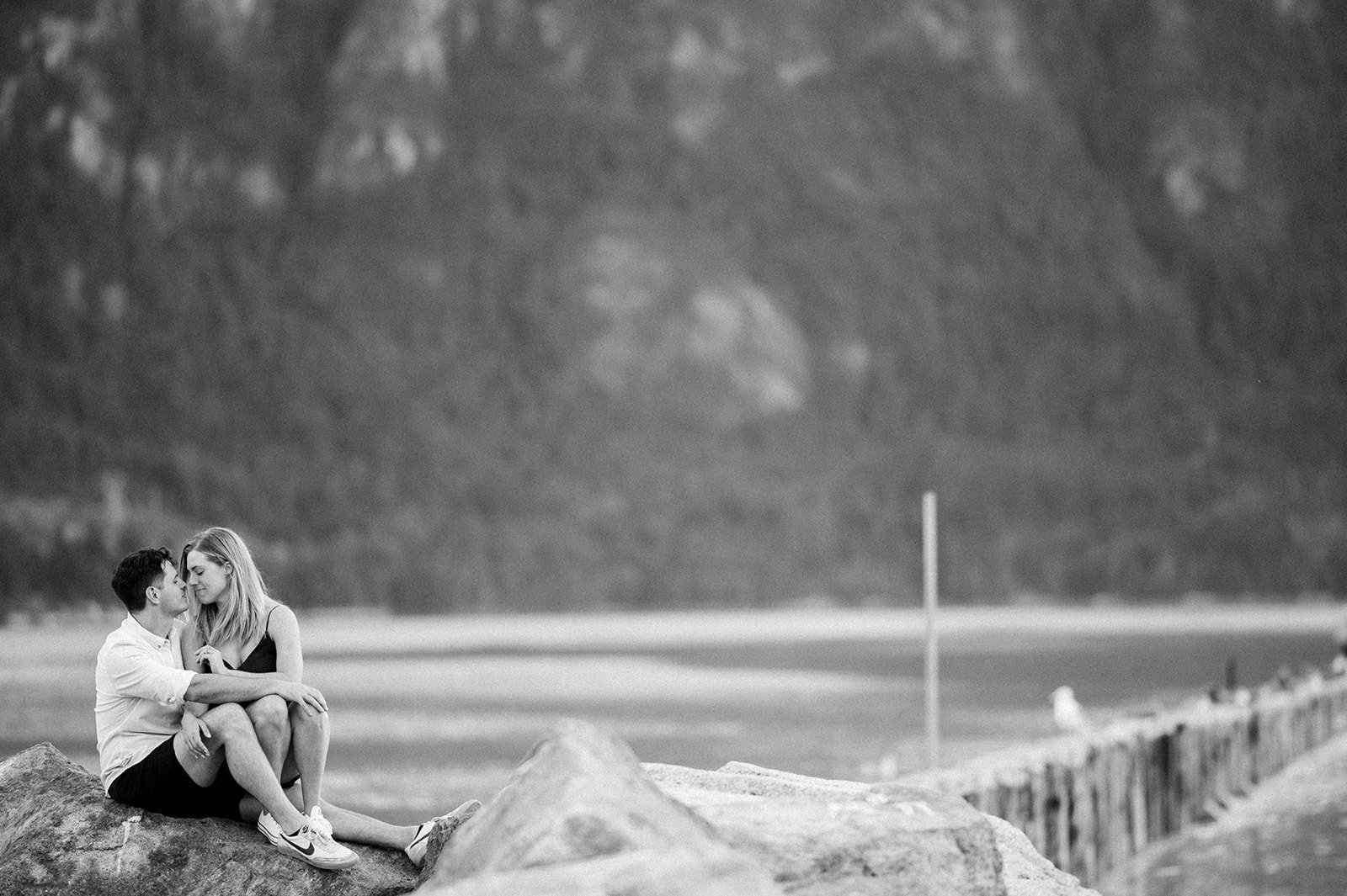 A blonde woman in a green dress sits on a man's lap in front of a beautiful green lake and mountain in Squamish, BC.