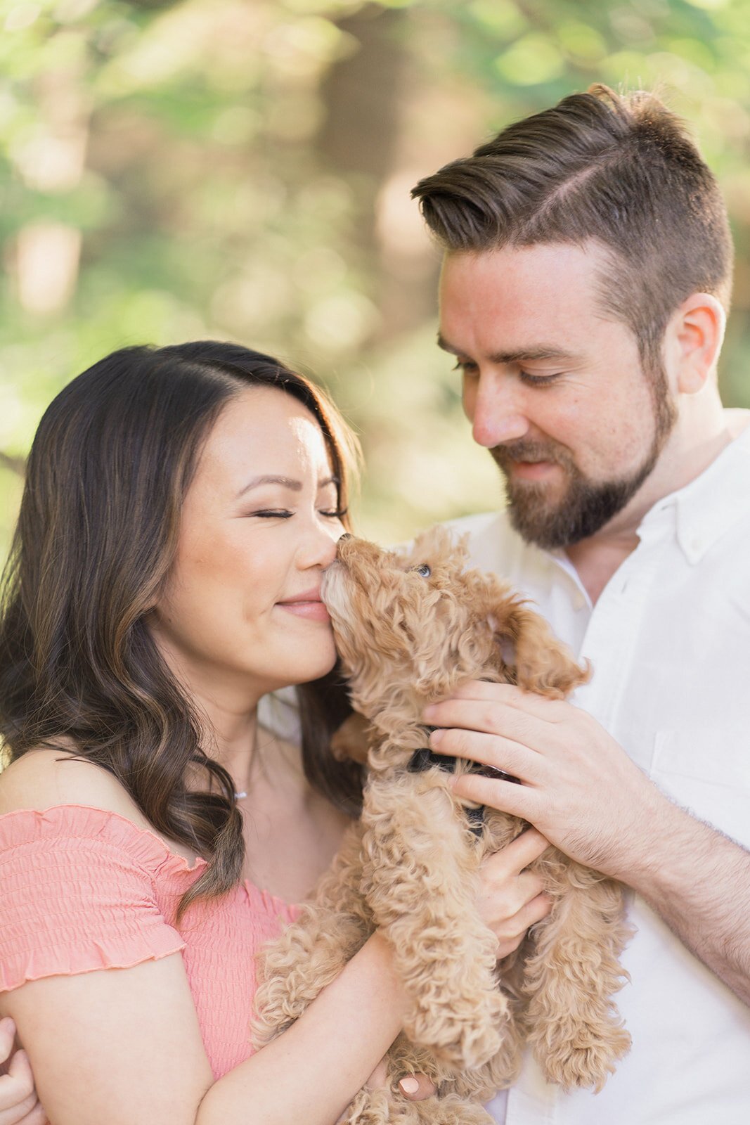 An adorable golden-doodle puppy lick's his moms nose as he is snuggled by her and his dog-dad.