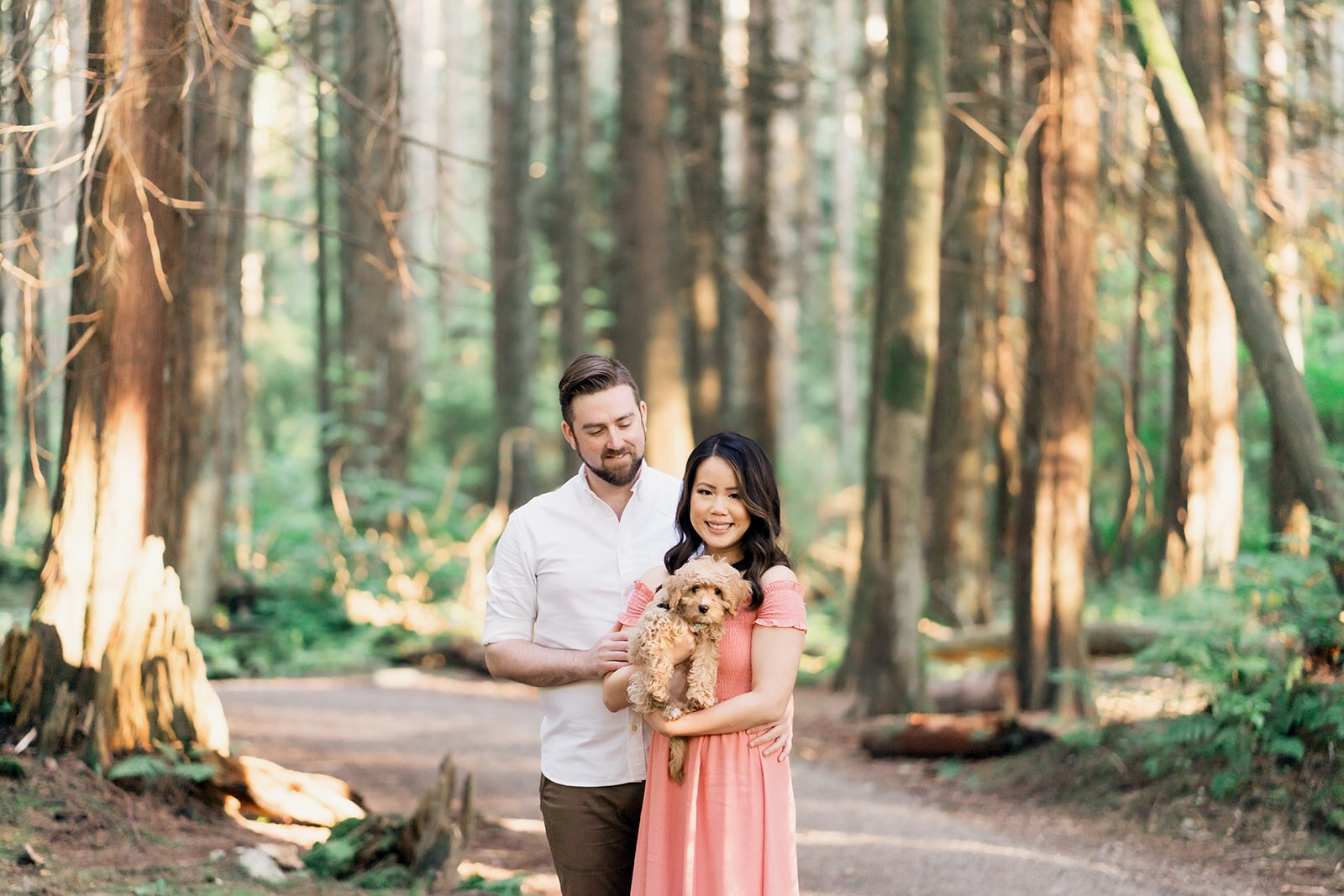 An engaged couple poses for the camera with their adorable curly-hair golden-doodle puppy in the forest of Stanley Park.