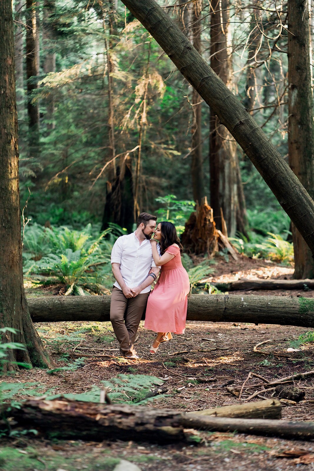 An engaged couple sits on a log in Stanley Park while cuddling. 