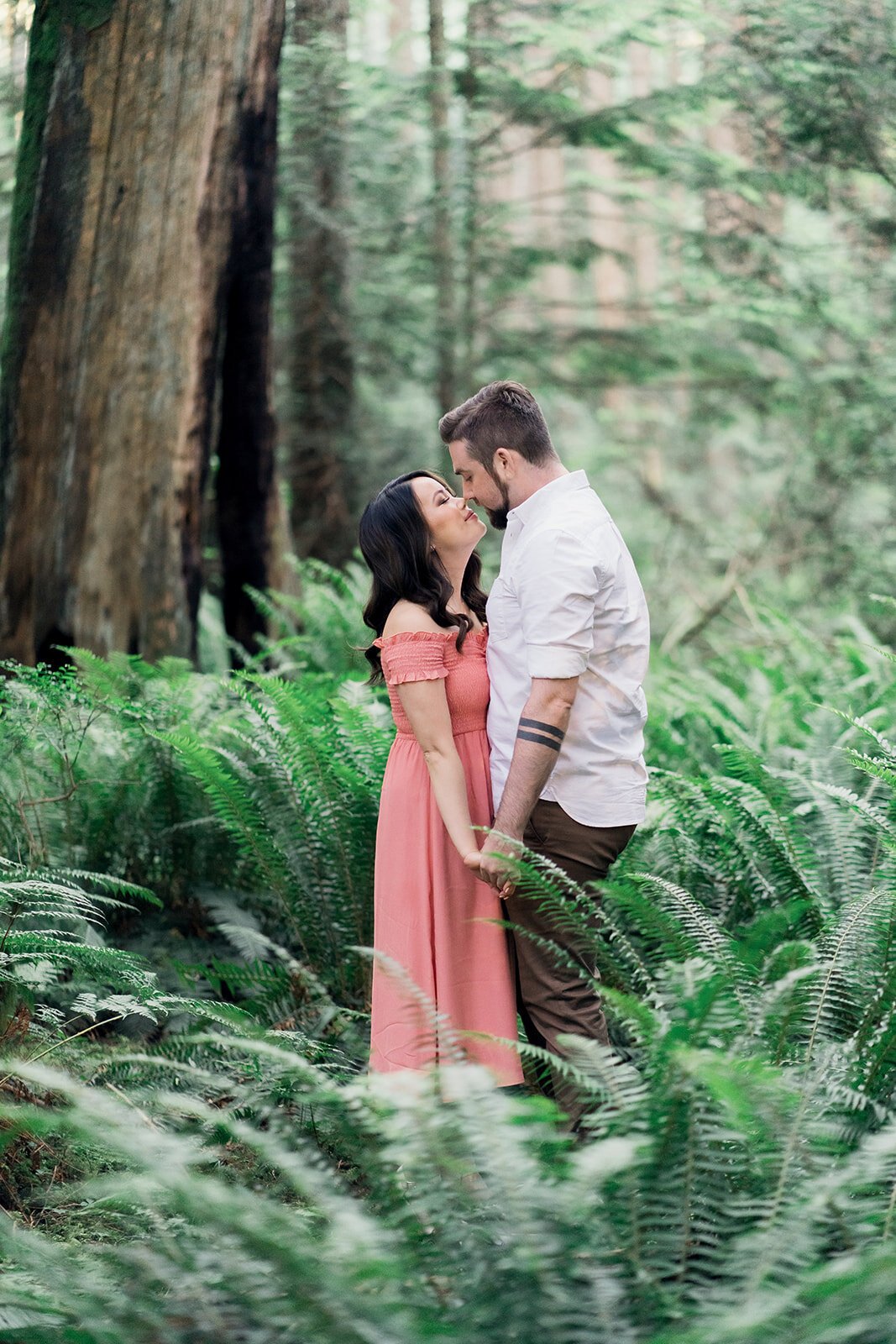 A woman in a cute pink dress holds her fiancee's hands as she kisses him, surrounded by ferns.