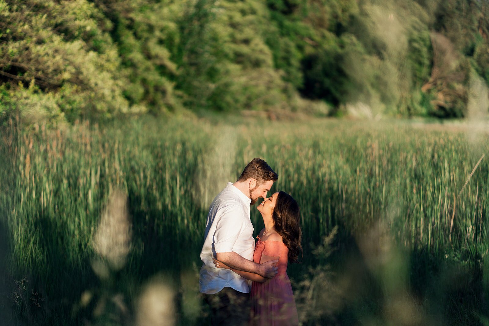 A woman in a pink linen dress holds her fiancee in a romantic grass field. 