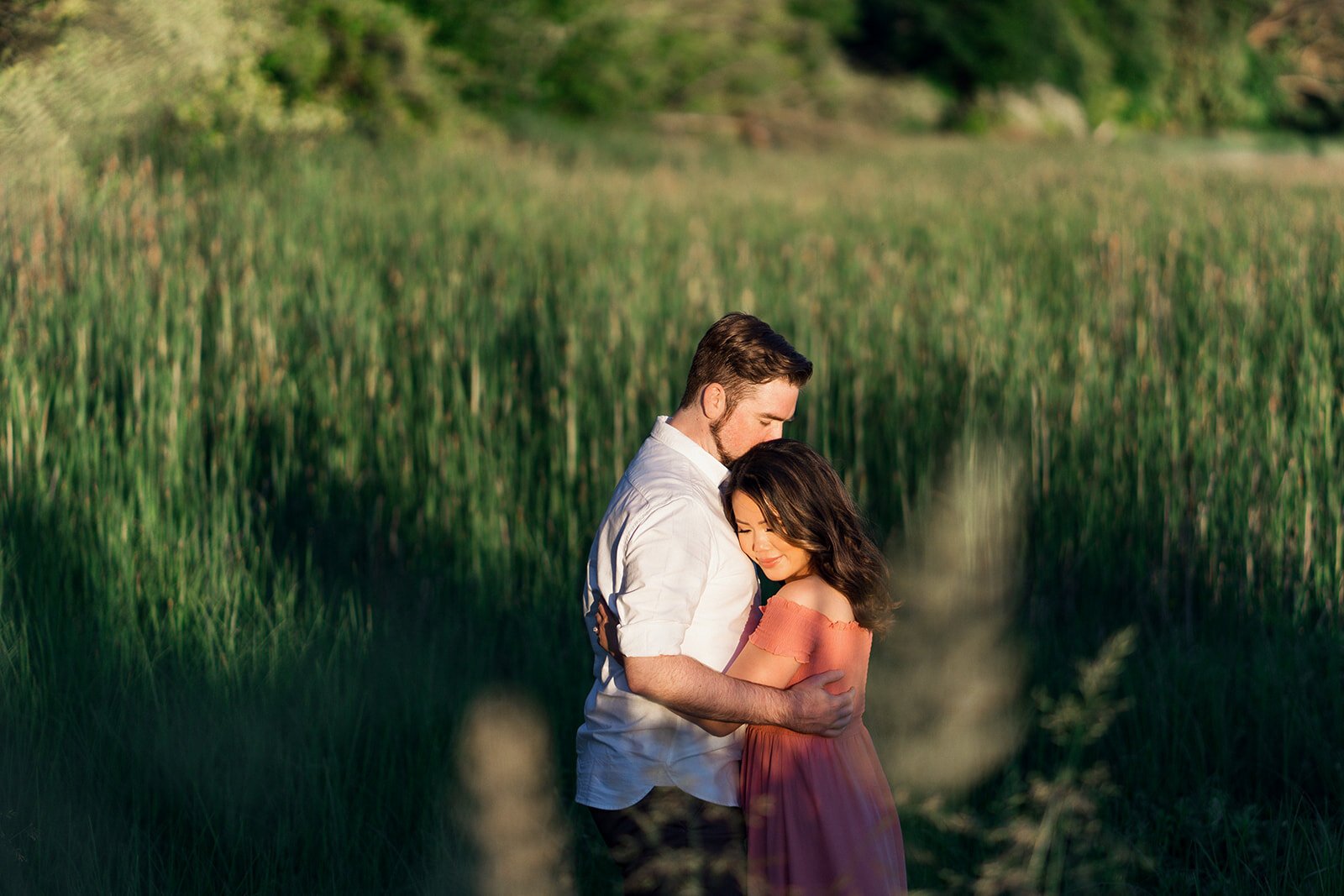 A woman in a pink linen dress holds her fiancee in a romantic grass field.