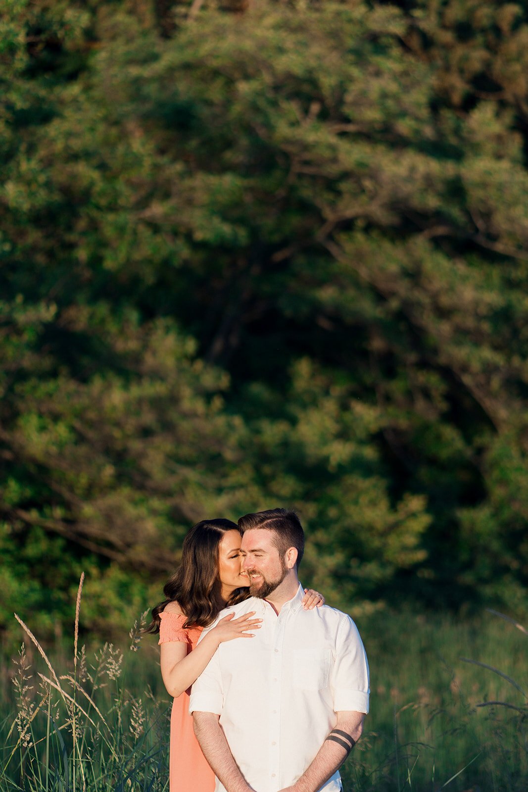 A cute woman in a pink dress hugs her fiancee from behind as they stand in a grass field surrounded by woods.