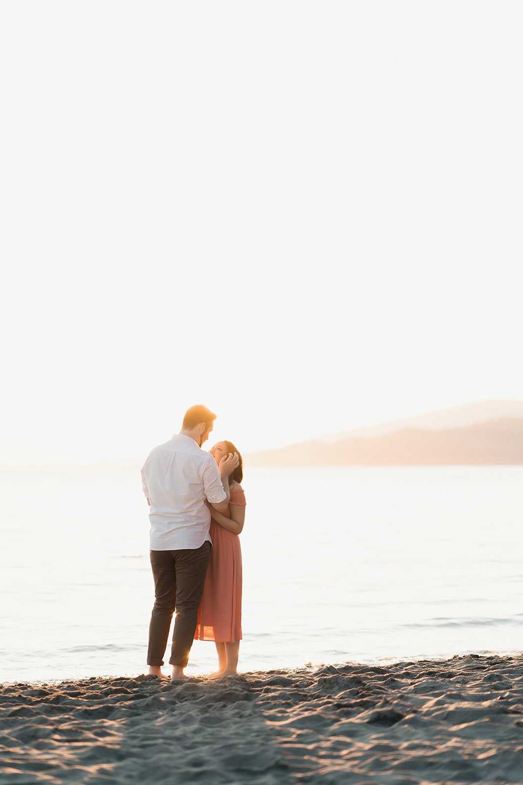 An engaged couple snuggle on a soft sandy beach lit by a gorgeous sunset in English Bay, Vancouver, BC.