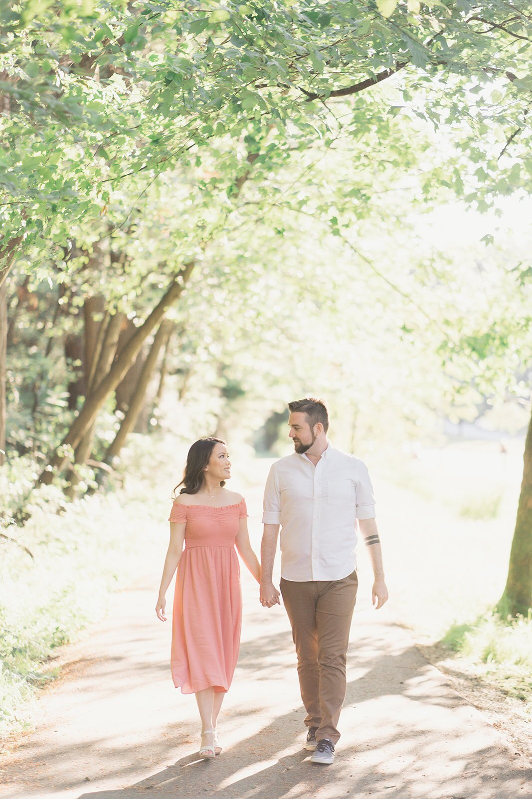 A beautiful woman in a pink dress holds her fiancee's hand as they stroll through the forest.