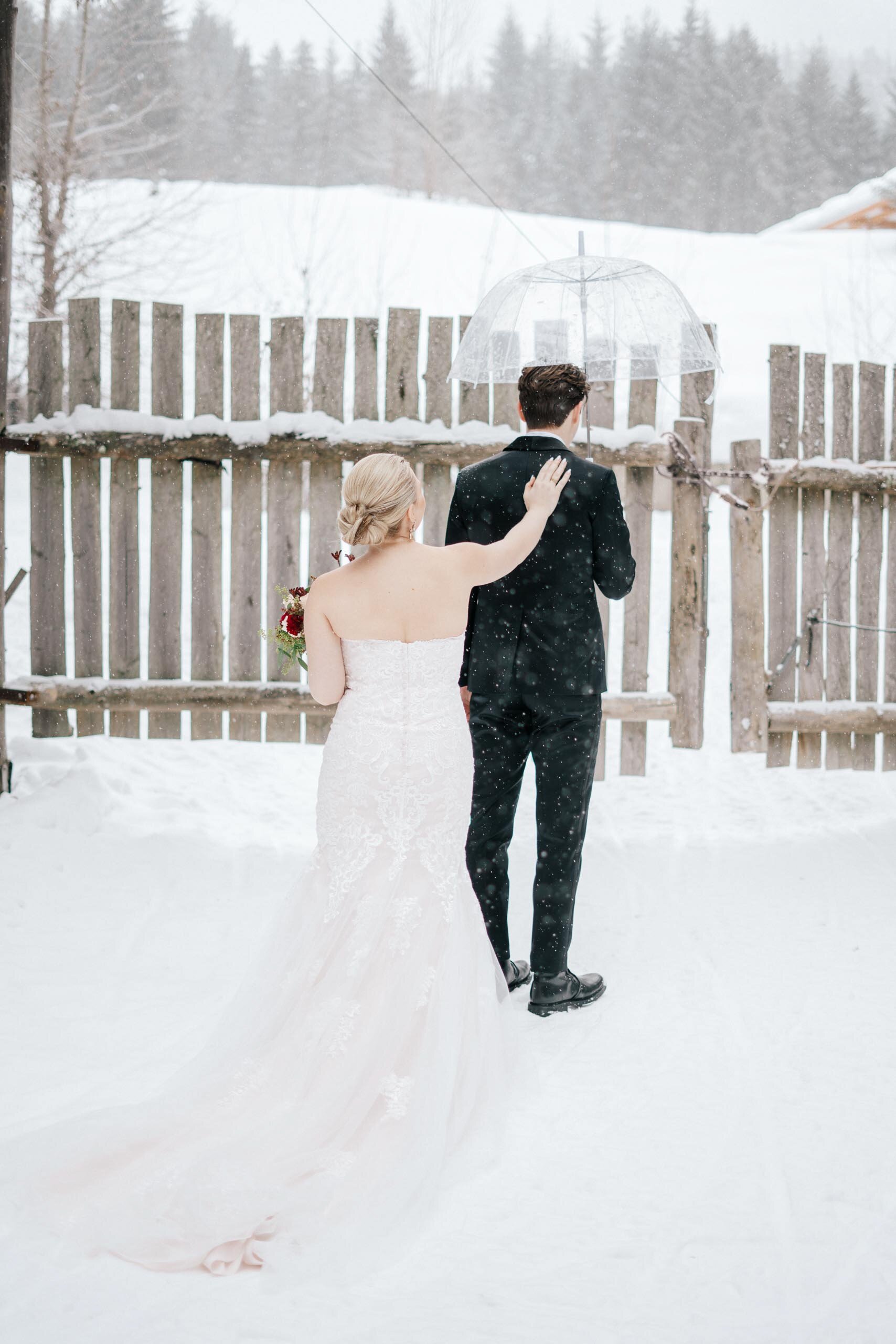 A bride in a long white dress with a lace train, taps her groom on the shoulder as the snow sprinkles. 