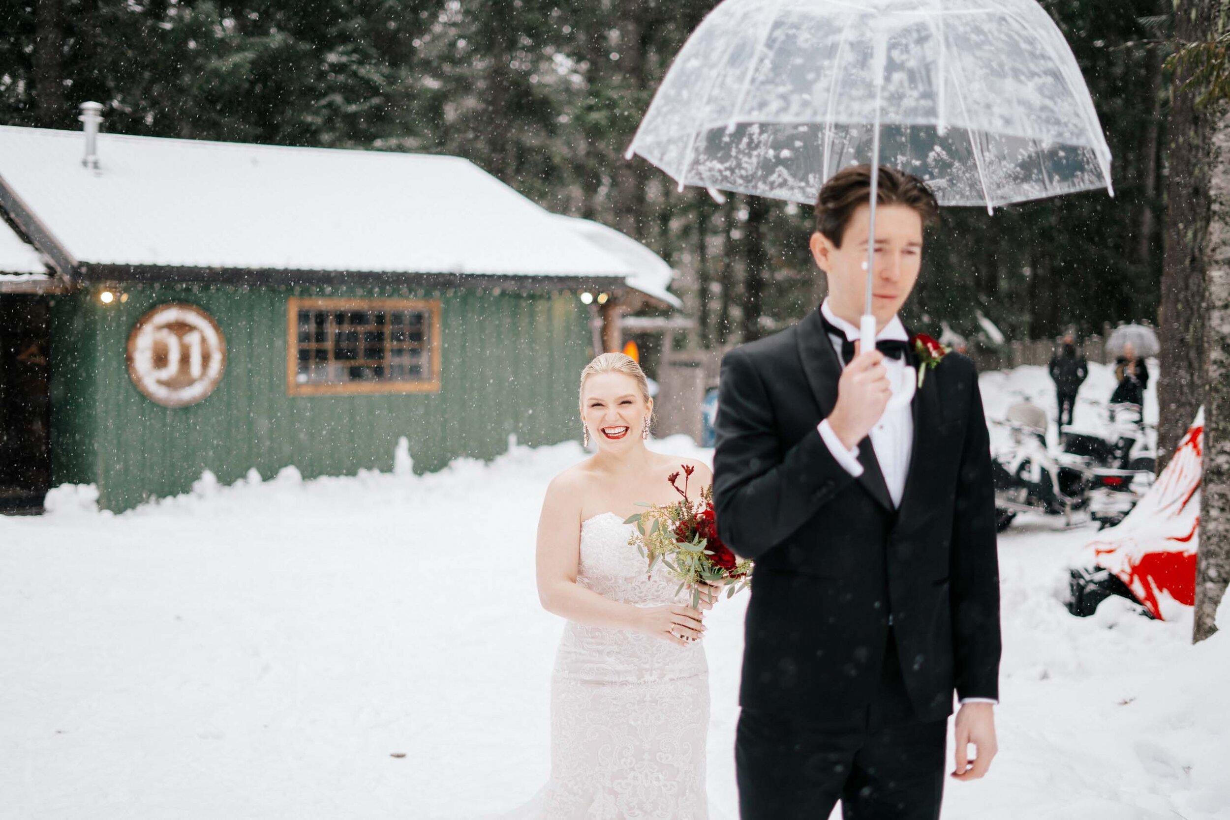 A bride stands behind her groom, before the first look, as snow falls on the couple.