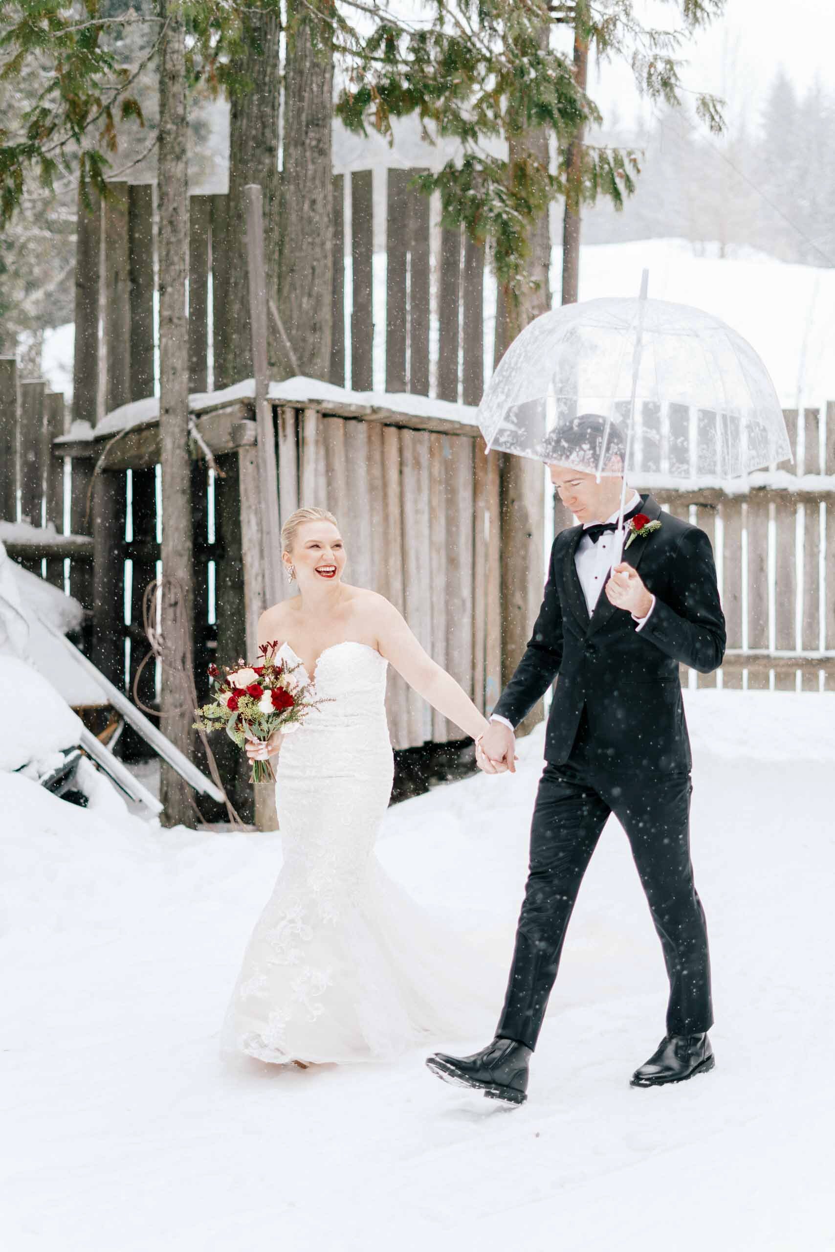 A bride and groom walk hand in hand, next to a wooden fence, through the snow in Whistler, BC.