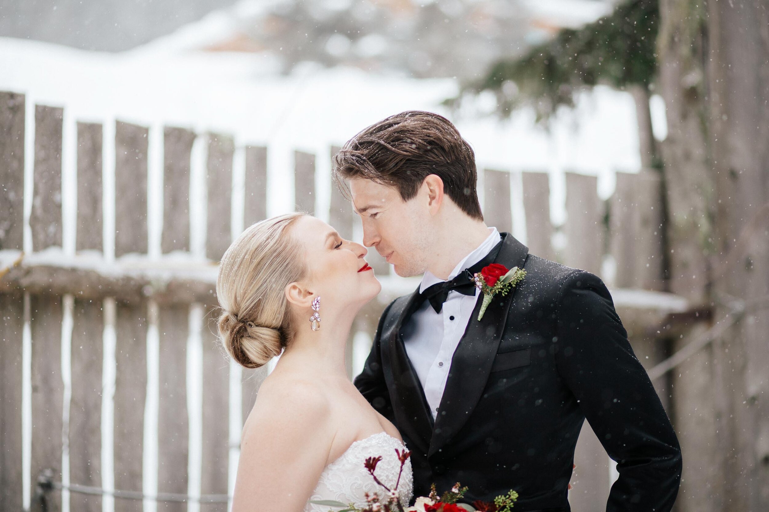 A beautiful blonde bride with bright red lips leans in for a kiss from her groom as the snow falls on them in Whistler, BC.