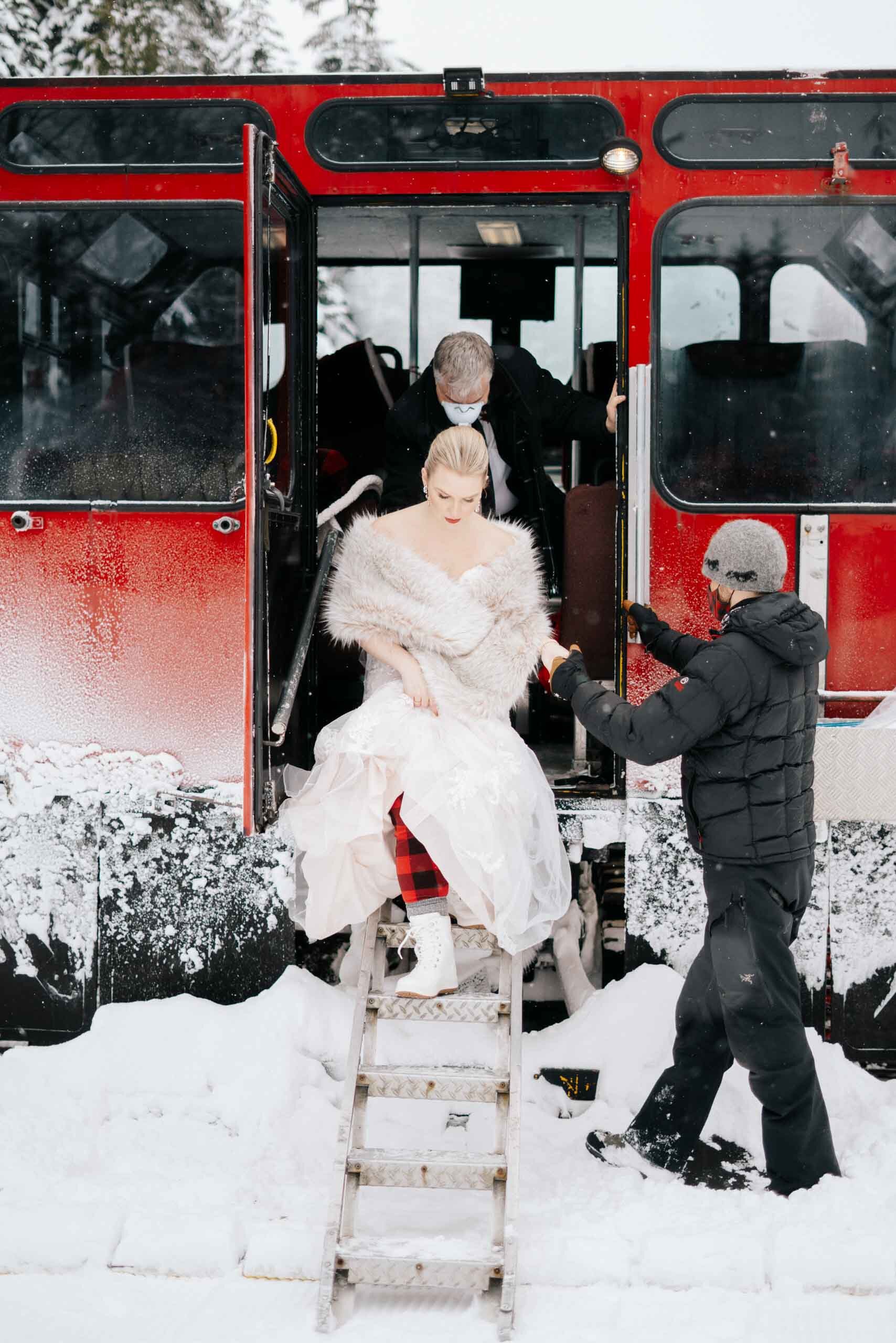 A bride in a lace dress and snow-boots disembarks from a red snow-mobile bus in Whistler BC. 