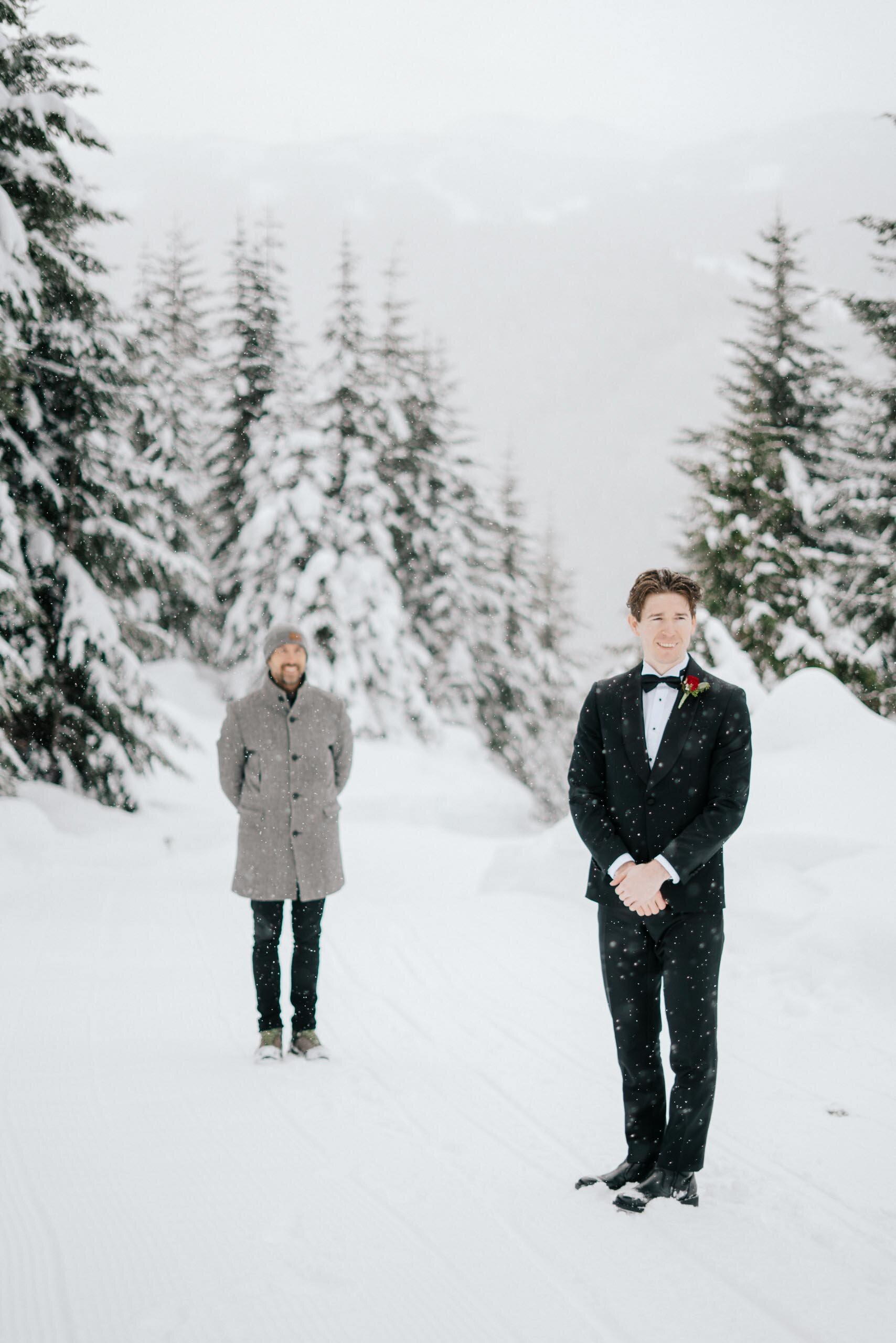 A groom waits for his bride in the snow, among snowy covered evergreens in Whistler BC.