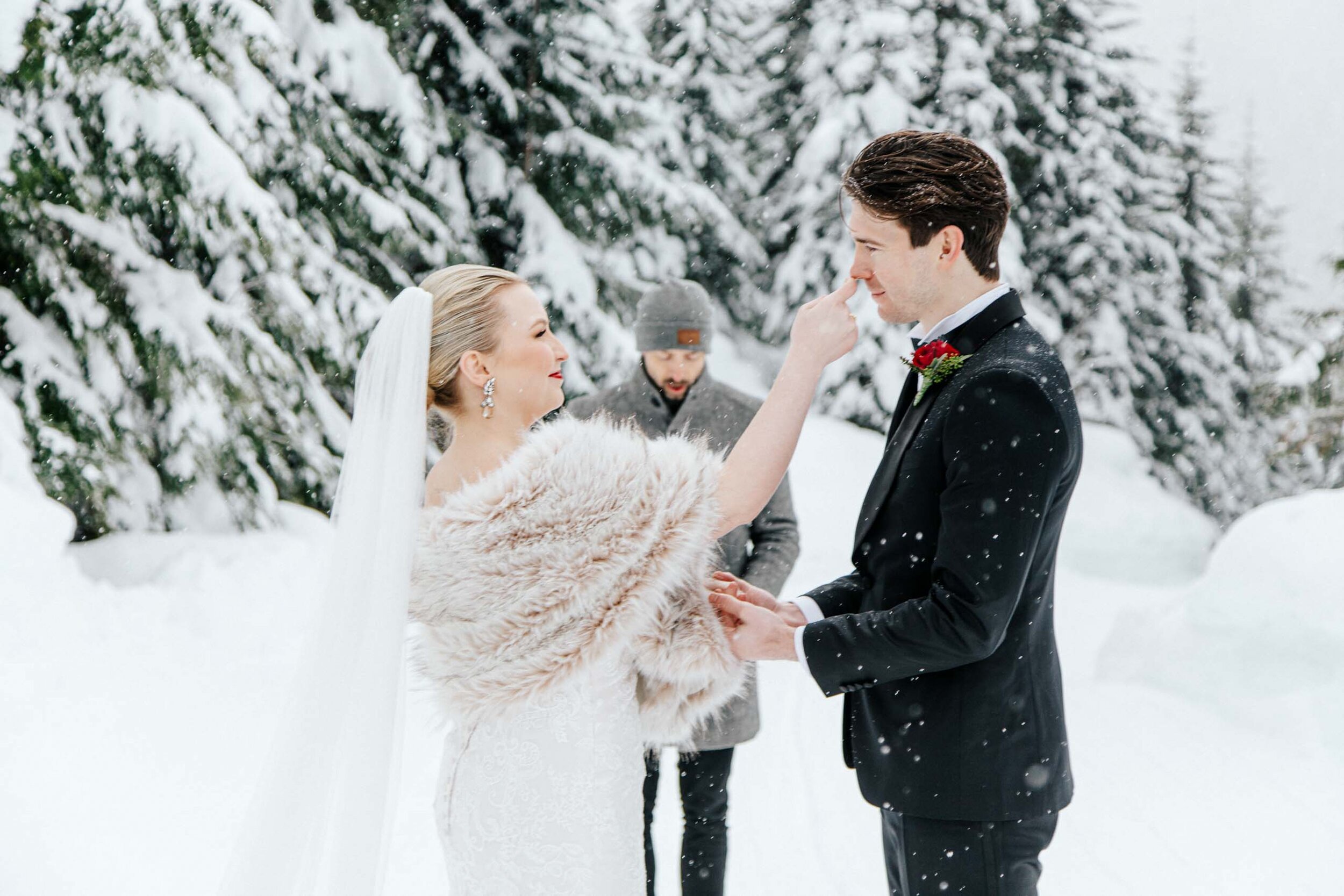 A snowy bride wipes her grooms nose as the snow drifts around them during their small ceremony in Whistler, BC.