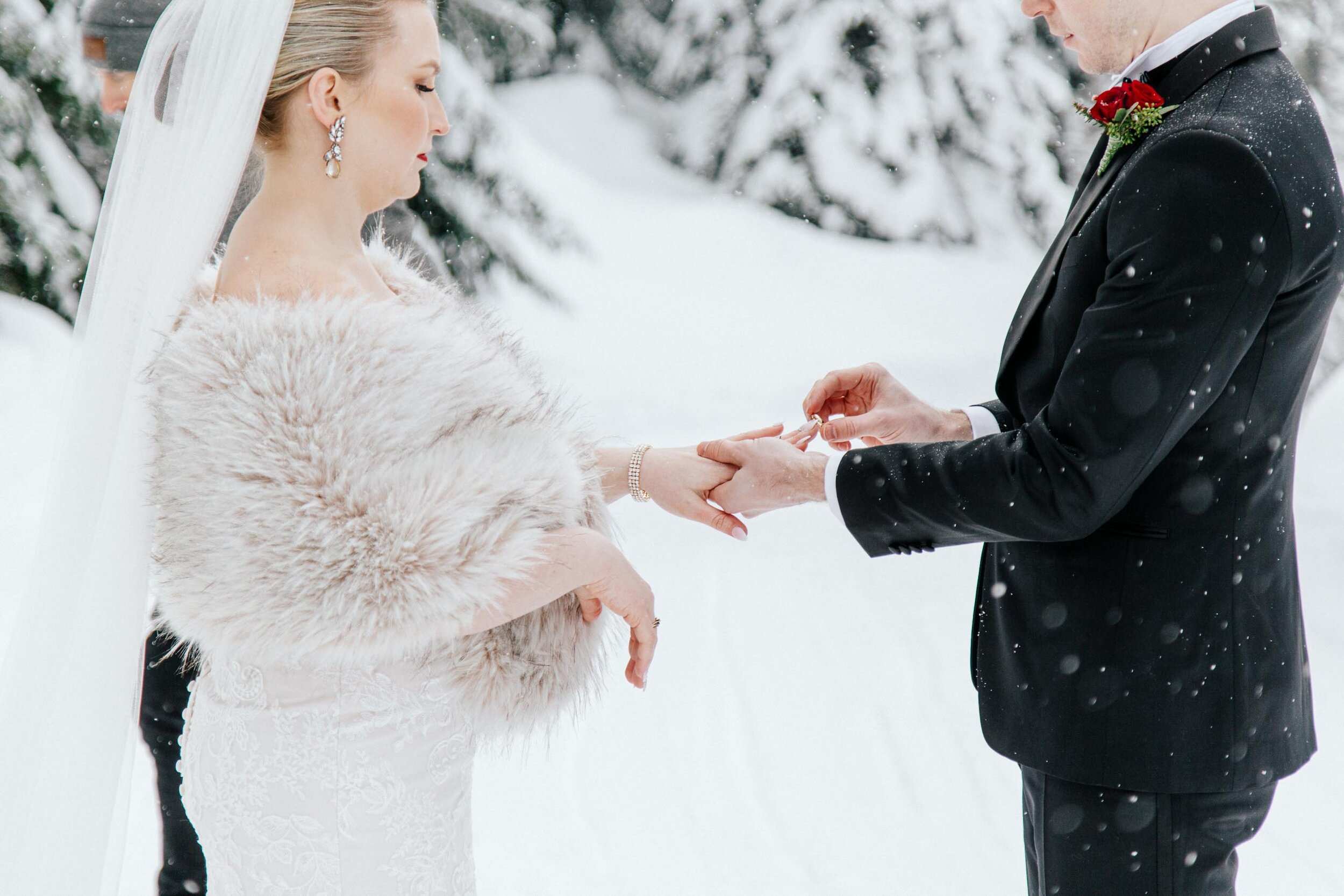 A groom places a ring on his brides finger as large snow flakes drift around them. 